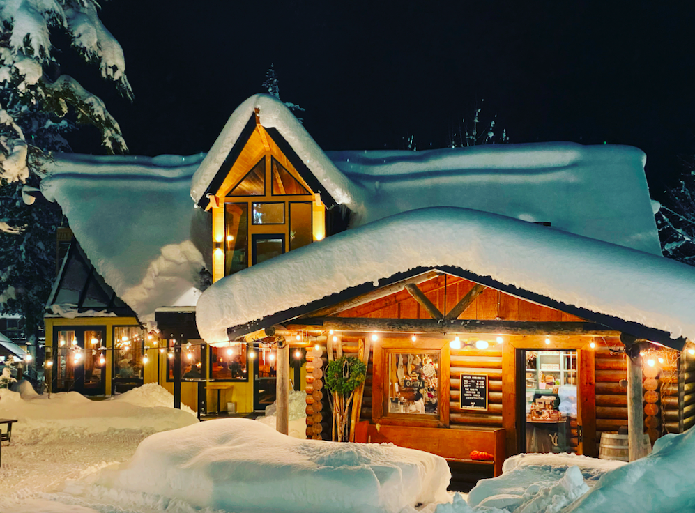 A snow-covered log building with warm interior lights, situated in a wintery forest scene at night. Mother Hubbard's Cupboard, base of Mt. Alyeska. Girdwood, Alaska. Jack Sprat restaurant celebrating 25 years.