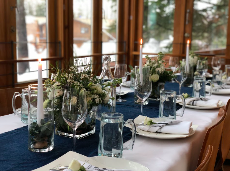 Formal dining table set with white tablecloth, blue table runner, white napkins, glassware, and floral centerpieces, inside a room with large windows and wooden walls.