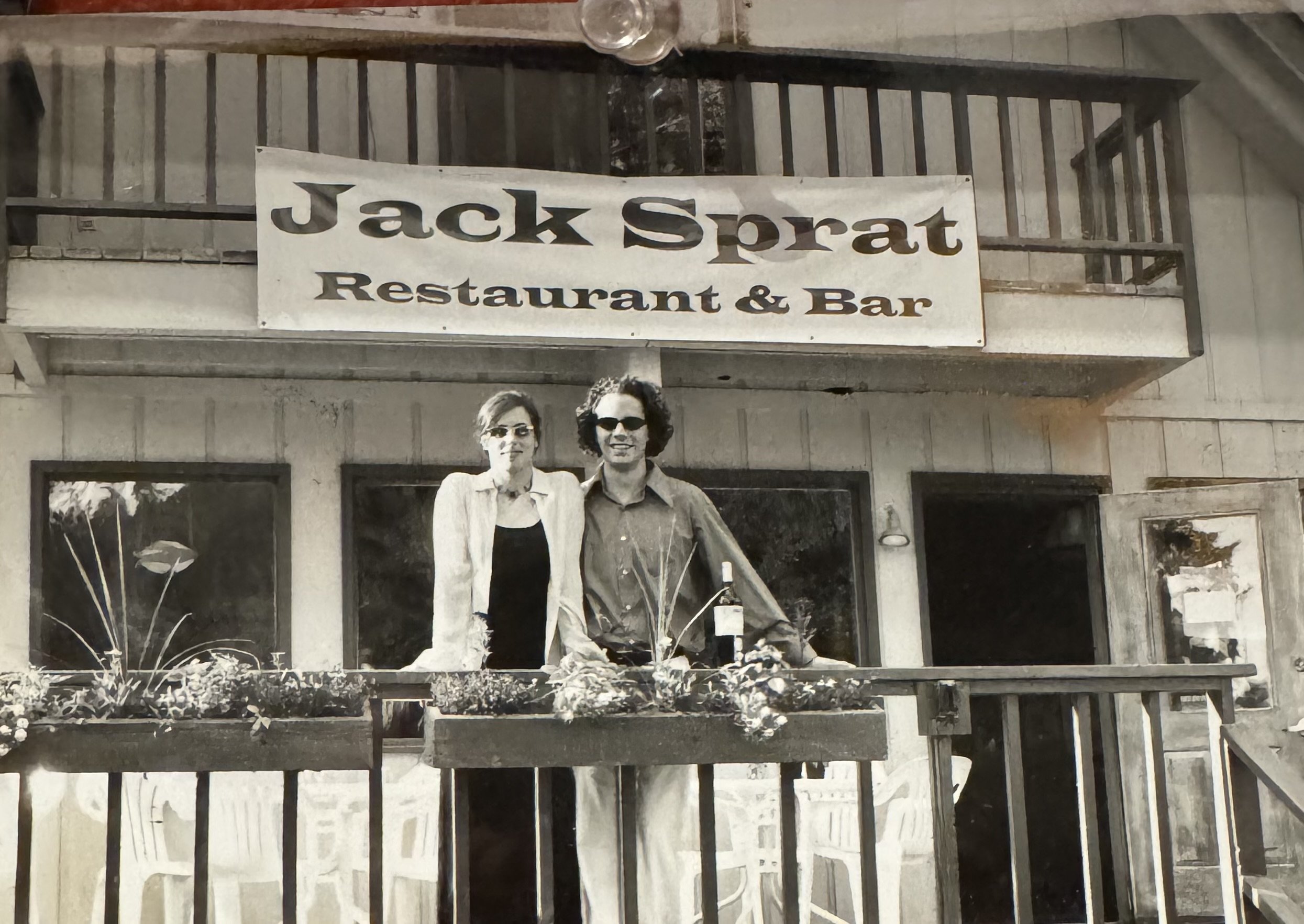 A black-and-white photo of two people, a woman and a man, standing together on a restaurant porch with flower boxes. They are smiling and wearing sunglasses. Above them, a sign reads "Jack Sprat Restaurant & Bar." Jen and Frans 
Girdwood, Alaska