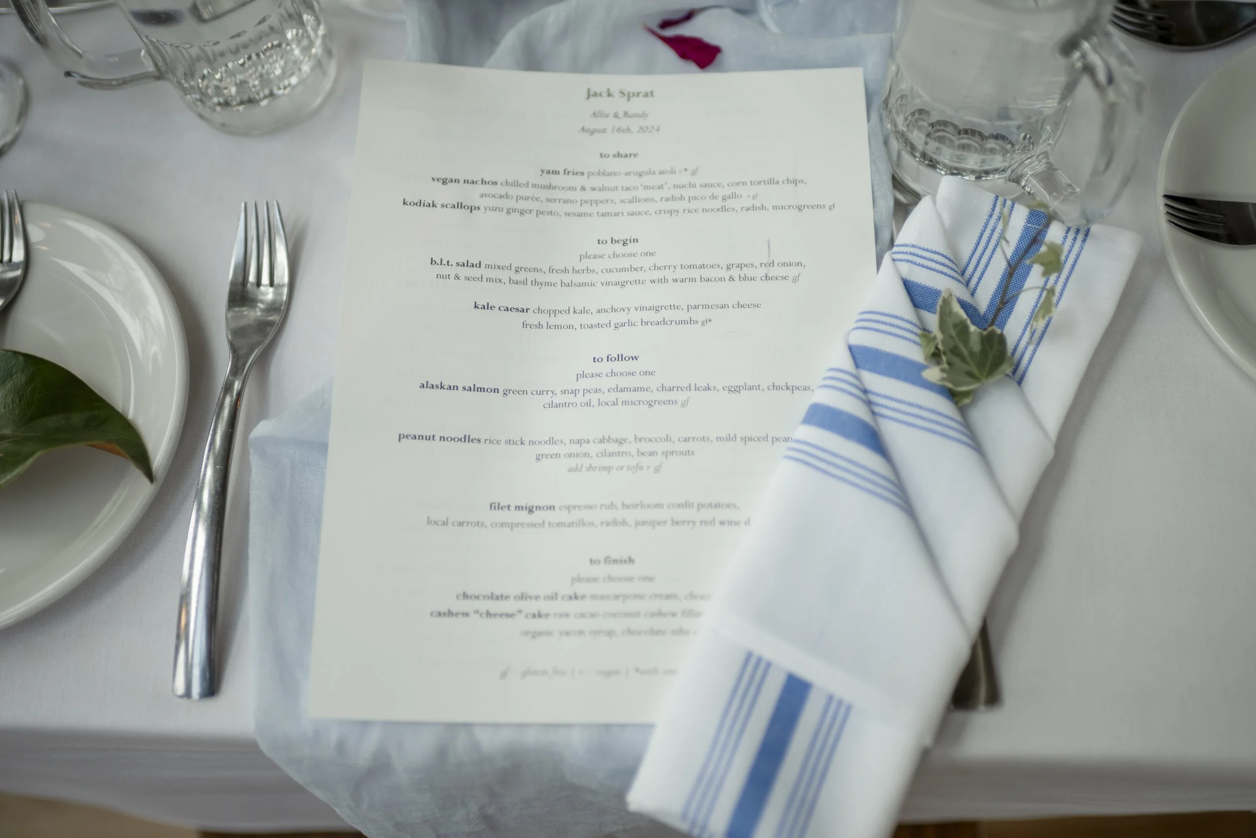Wedding table setting with a printed menu, a white napkin with blue stripes and a small decorative green plant, clear drinking glass, and silverware arranged on a white tablecloth.