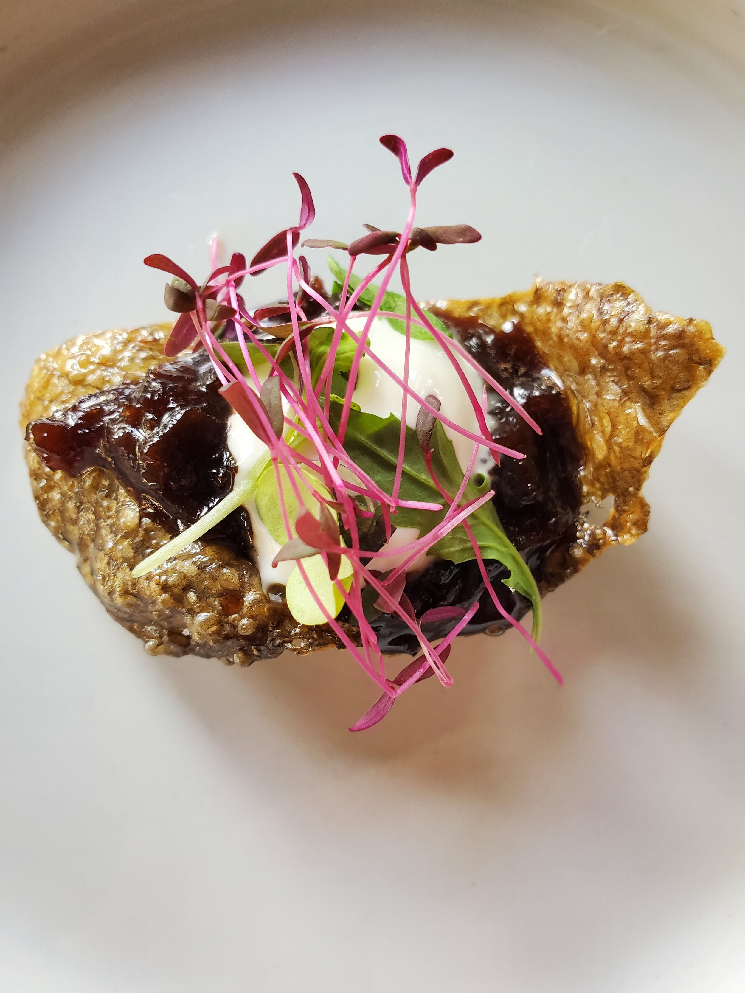 Close-up of a gourmet appetizer on a white plate, featuring a crispy rice cracker topped with dark sauce, microgreens, and edible flowers.