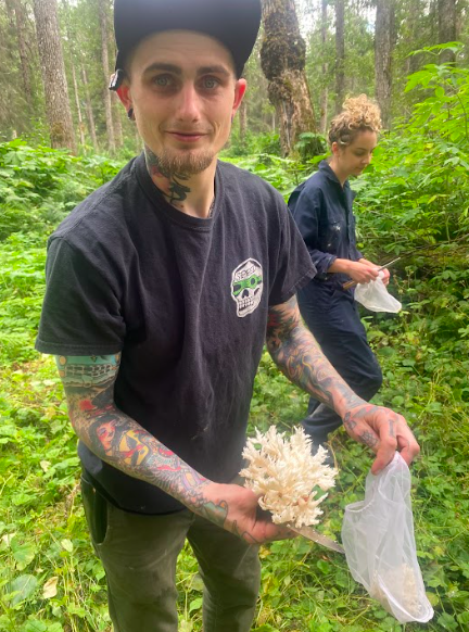 Man with tattoos holding a large white coral in a forest, woman in the background collecting something from plants. Foraging in Alaska. Forest to table. Alaska boreal forest. Chugach National Forest. Chefs that forage.