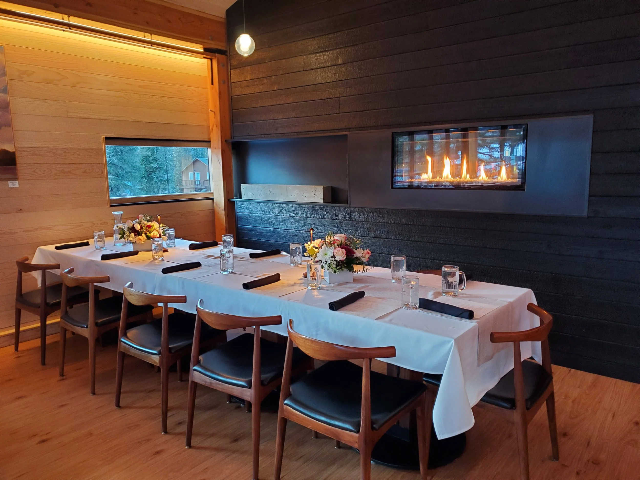 There are two floral centerpieces, and a wall-mounted fireplace with flames behind a glass panel. The room has wooden walls and floor, with a window showing the outdoors. Shou Sugi Ban wall.  Architecturally designed, hospitality design.