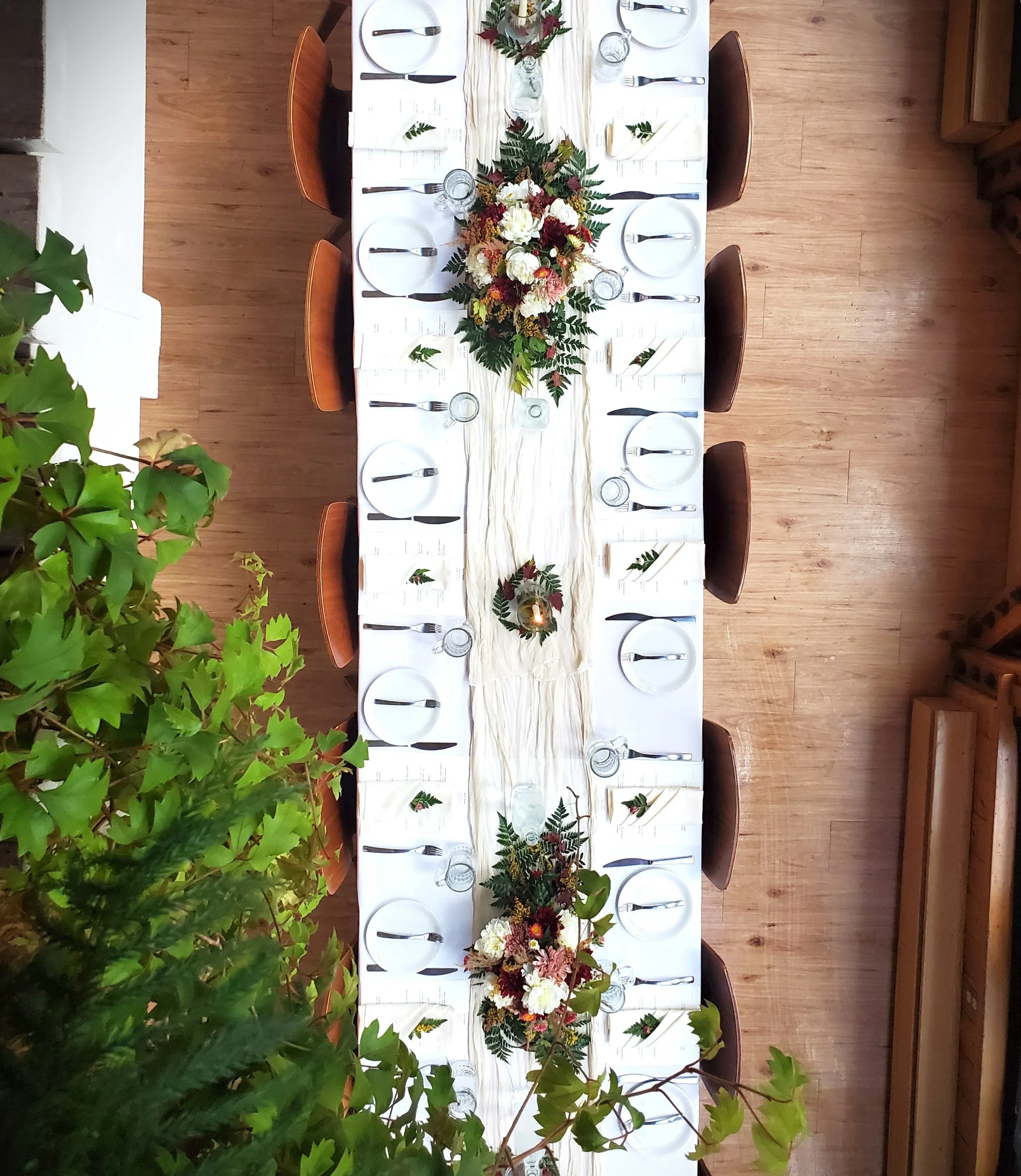 Long dining table set for a meal, decorated with floral centerpieces, white napkins, and glassware, with chairs on each side and greenery in the foreground. Bridal wedding table. private event table. Rehearsal dinner. Classy Alaska wedding