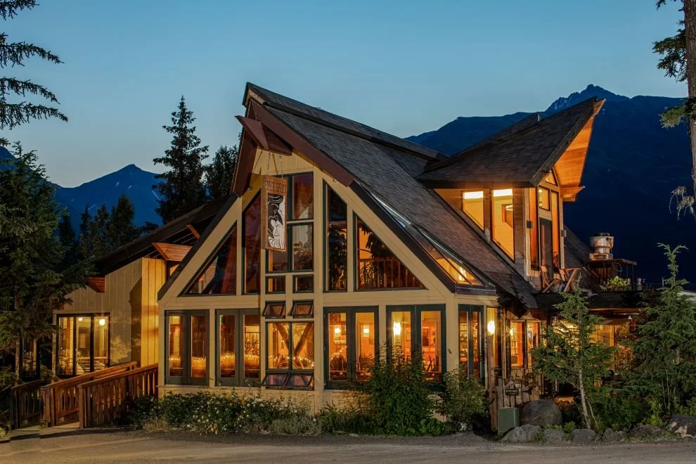 A modern designer stand alone restaurant with large glass windows and a geometric roof. Architecturally illuminated from inside, situated at the base of Mt. Alyeska surrounded by trees at dusk.