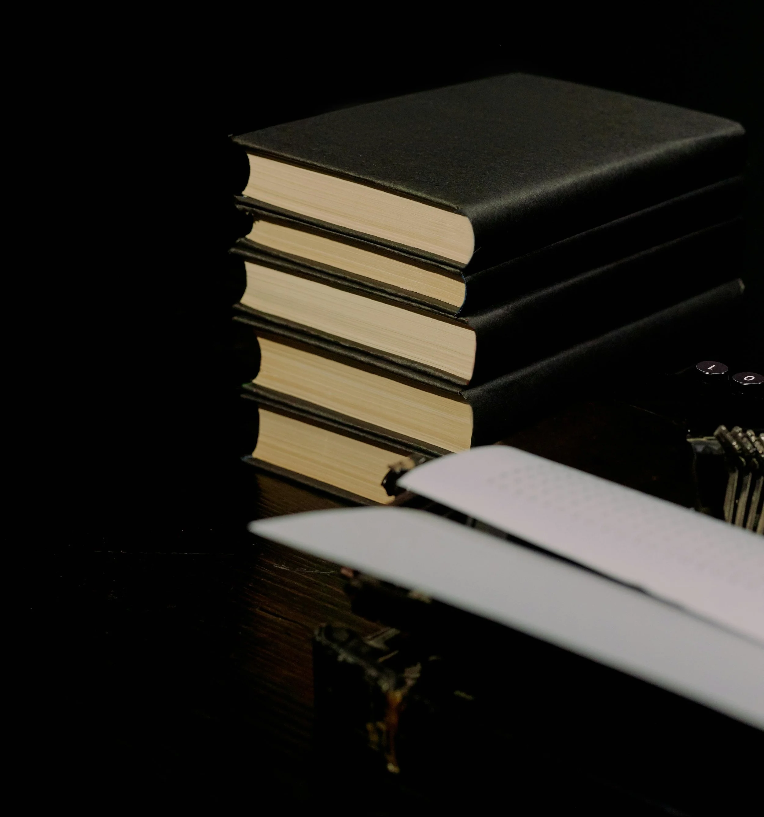 Stack of five black hardcover books with beige pages, partially illuminated on dark wooden surface, with a white shelf and small electronic device in the background on the Ruminous website.
