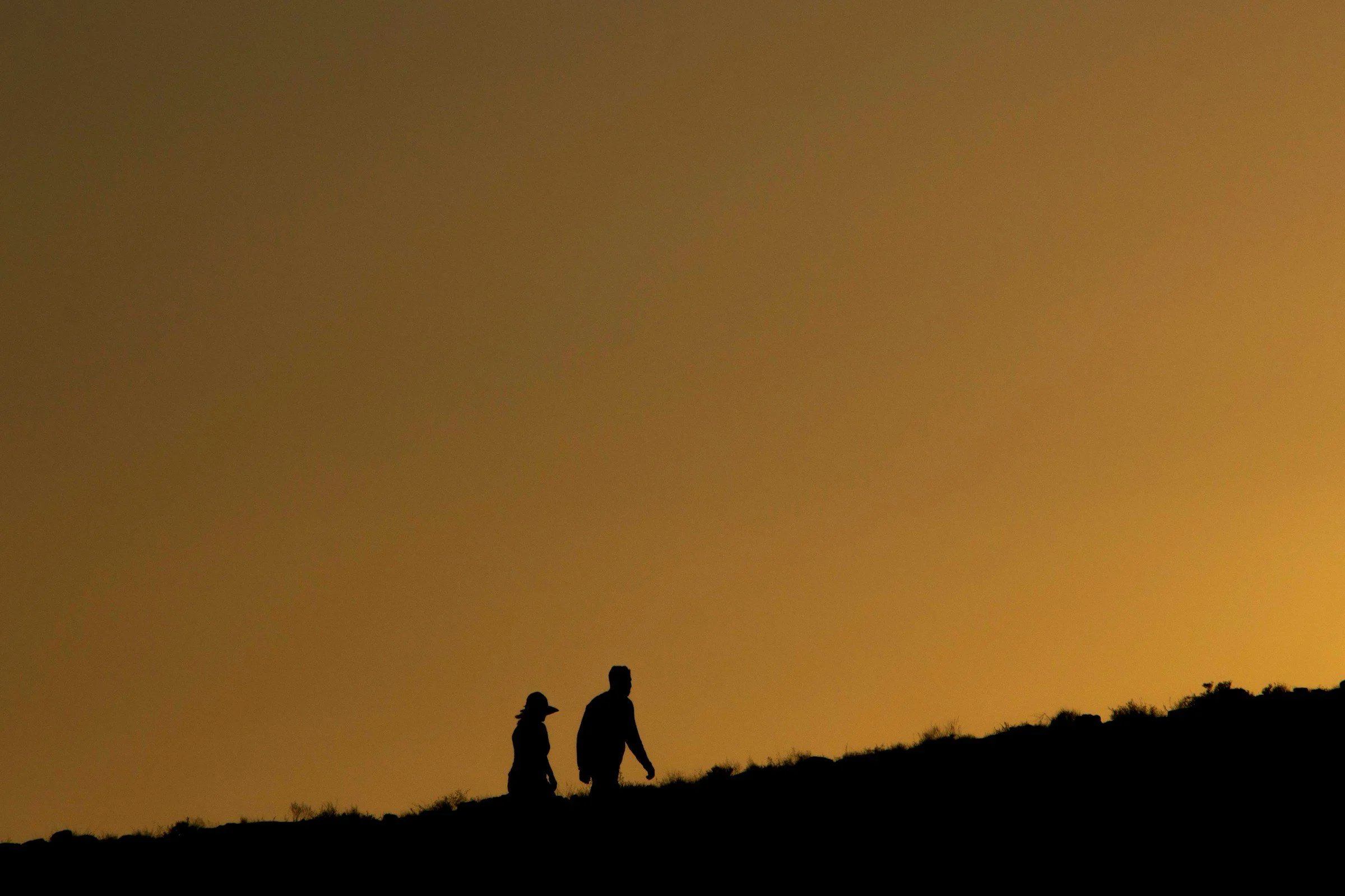Silhouettes of two people walking on a hill at sunset or sunrise with a warm orange sky.