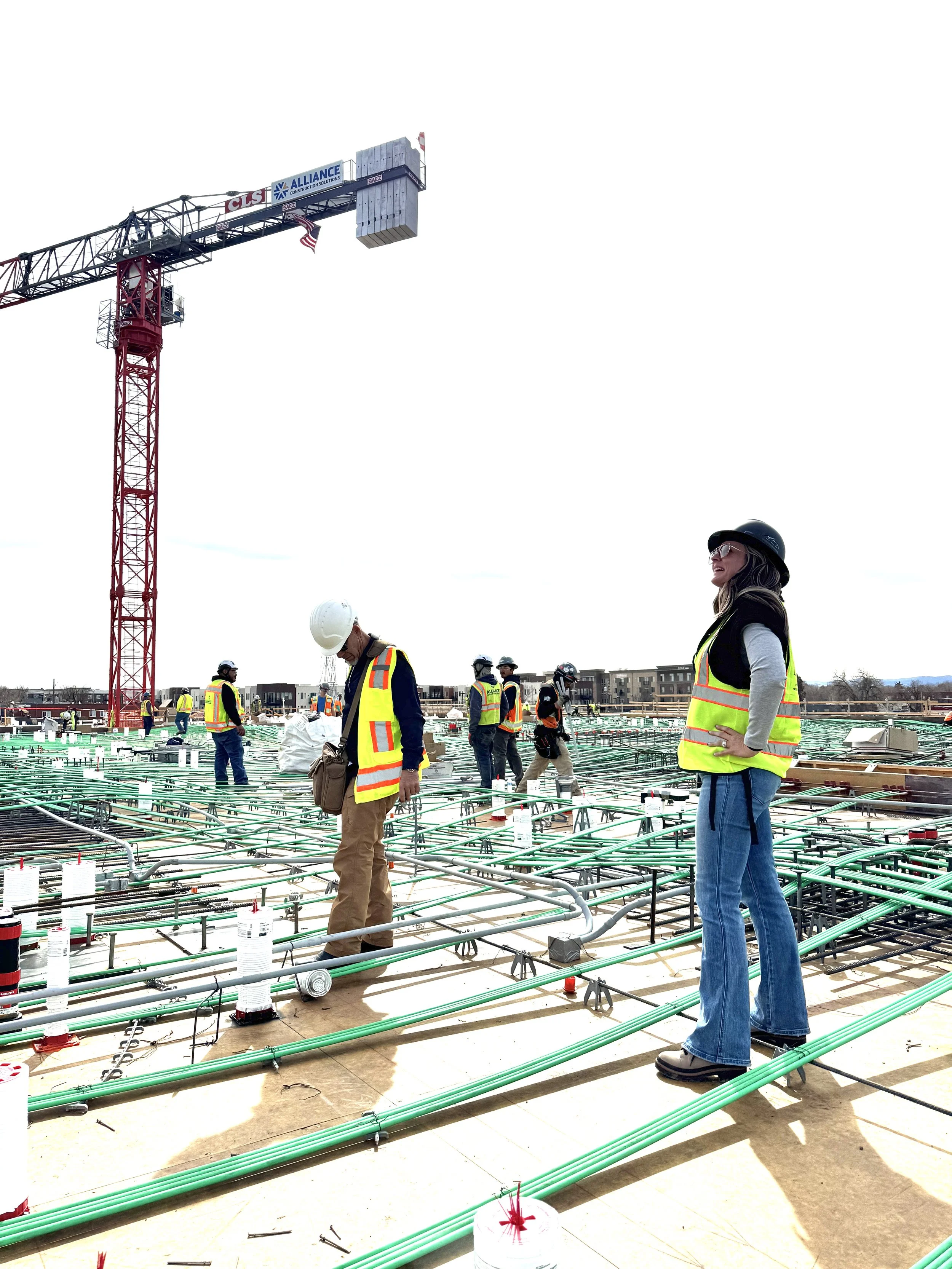 Construction workers with safety vests and helmets on a building site, featuring a large crane overhead and a framework of pipes and materials.