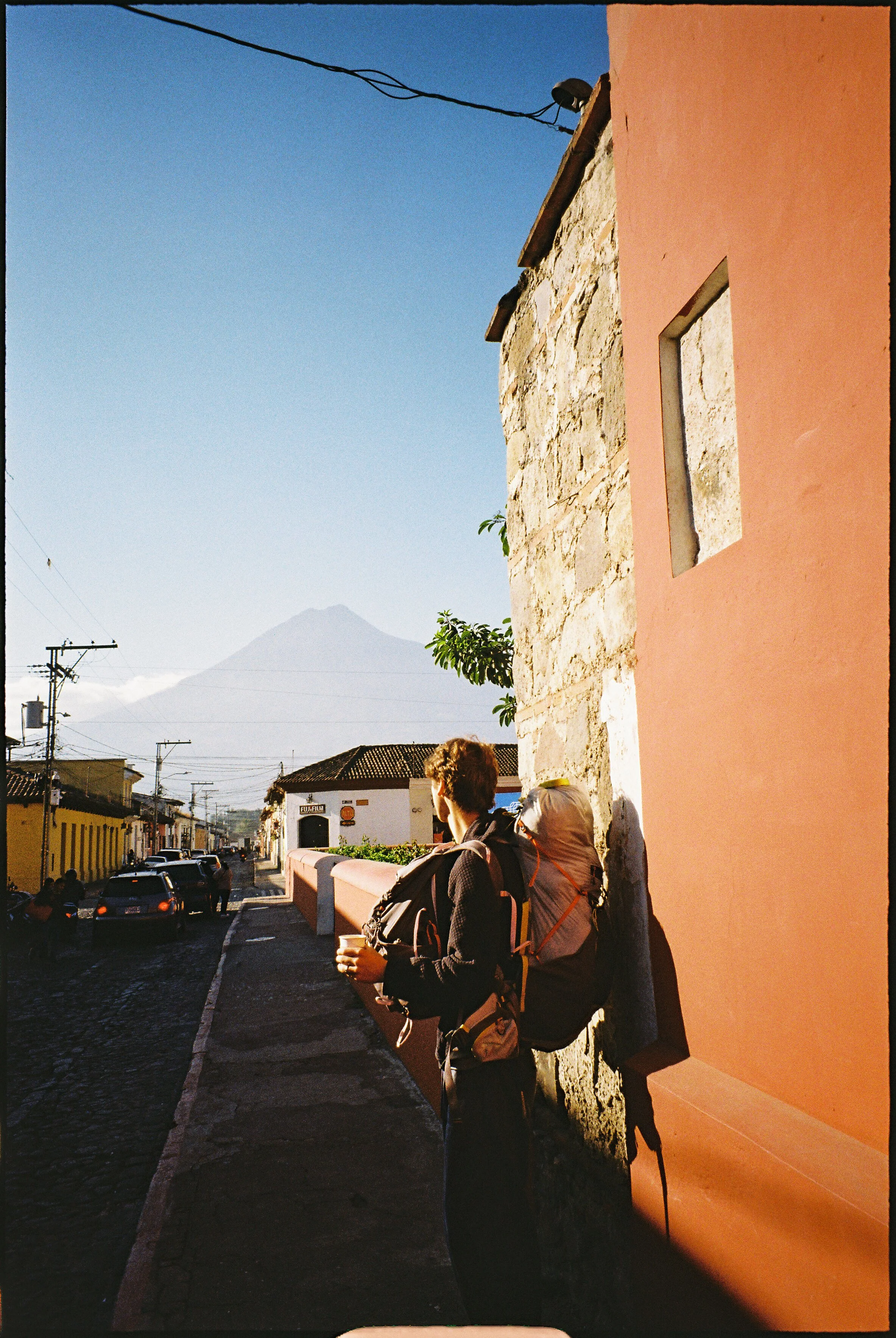Person with a backpack standing on a sidewalk in a colorful town, with mountains in the background.