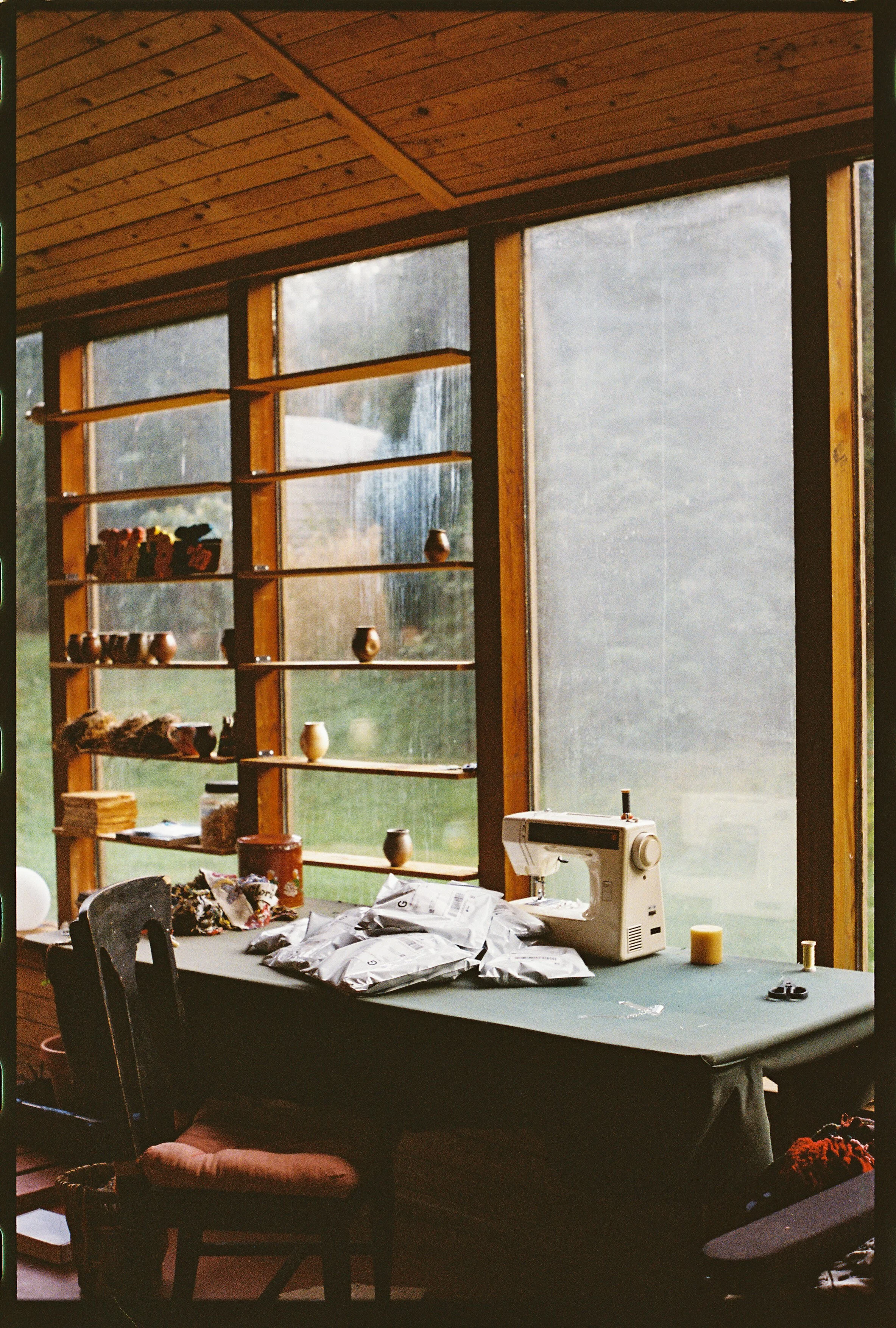 Interior of a rustic room with large windows and wooden walls. A sewing machine sits on a table surrounded by fabric, scissors, and other sewing supplies. Shelves hold pottery and decorative items, with an outdoor view visible through the window.