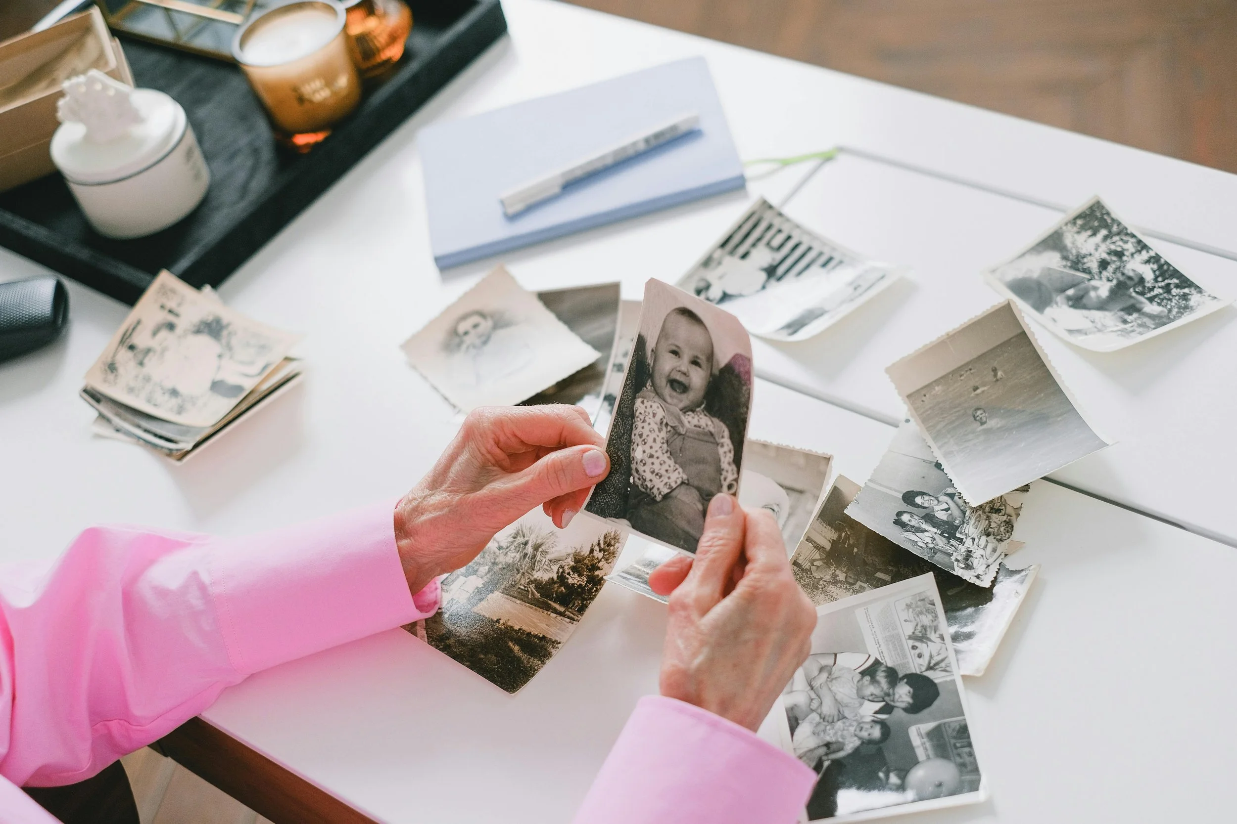 Two hands are holding a photo of a smiling baby while other  black and white photographs are on the tabletop all slightly curved due to age while being organized sparking memories and reminiscing by the owner