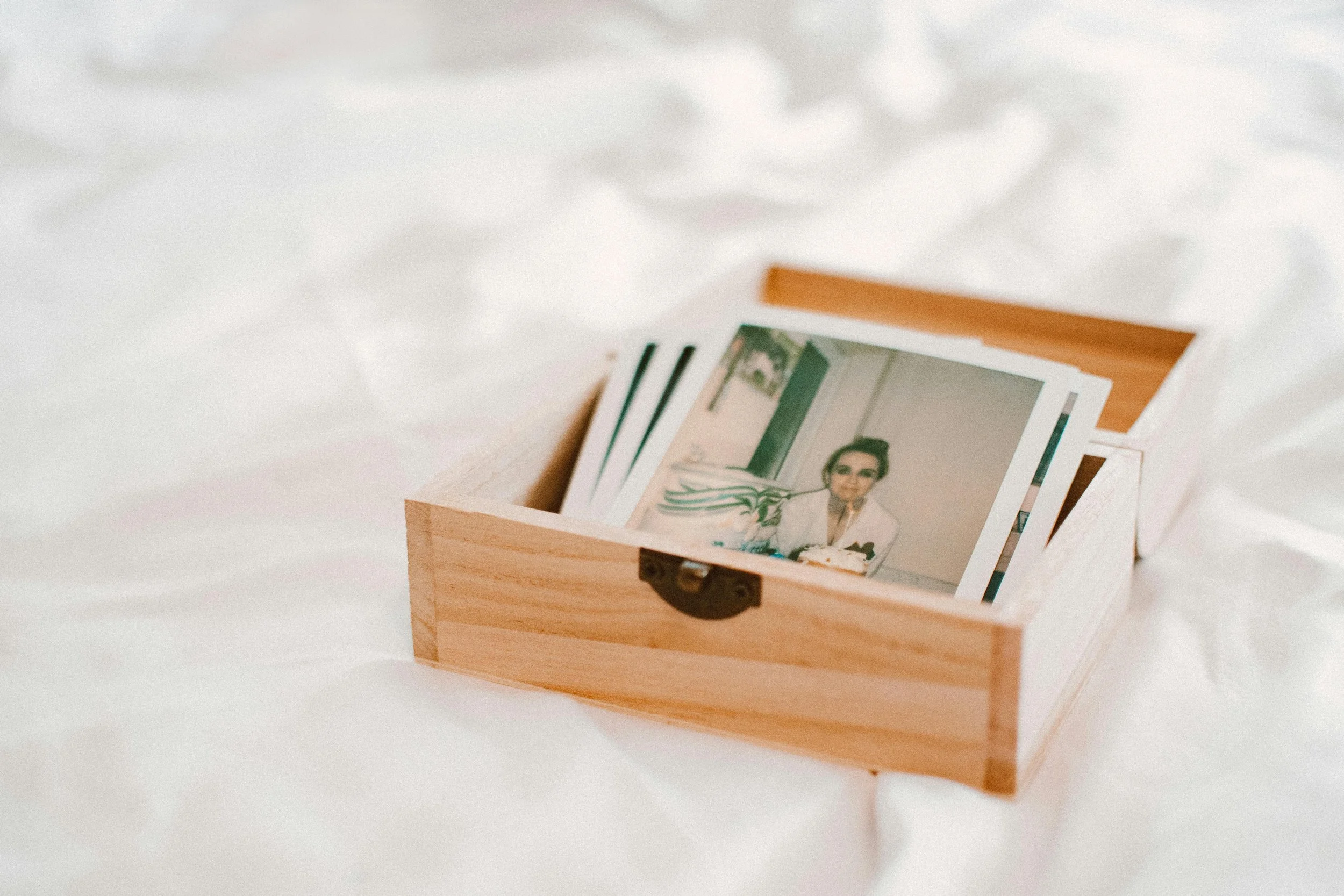 A light wooden box with a latch, containing printed photographs, resting on a white fabric surface.