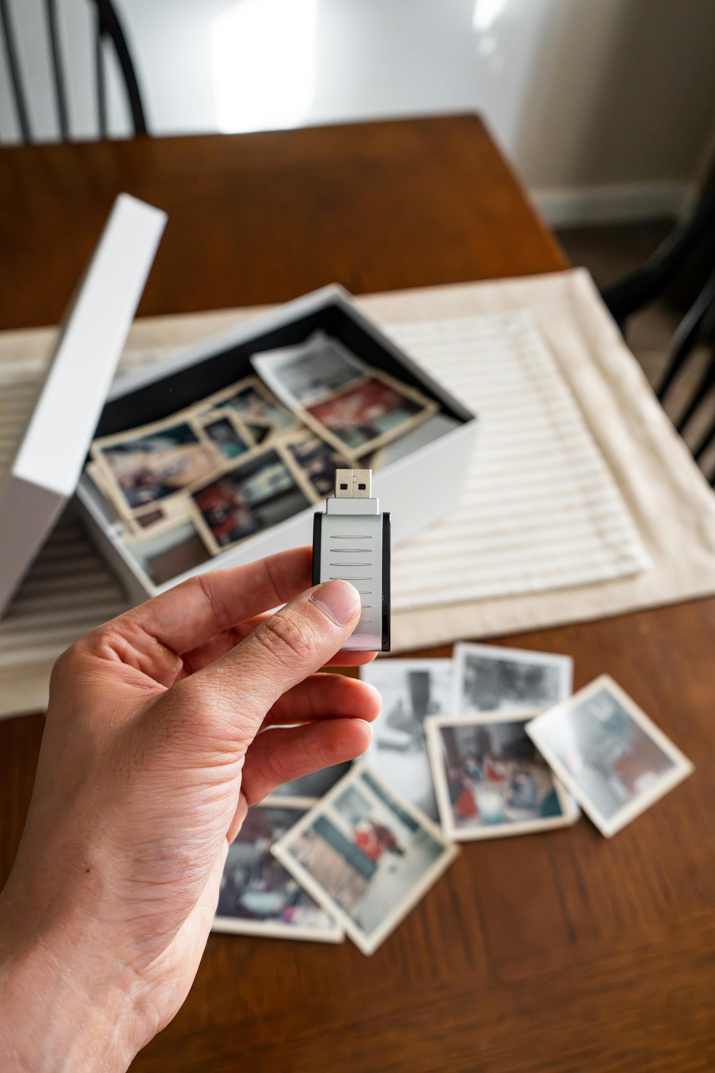 A flash or jump drive is being held while a dining room table is in the background with older photos on the table and more photographs in a shoe box or archival box