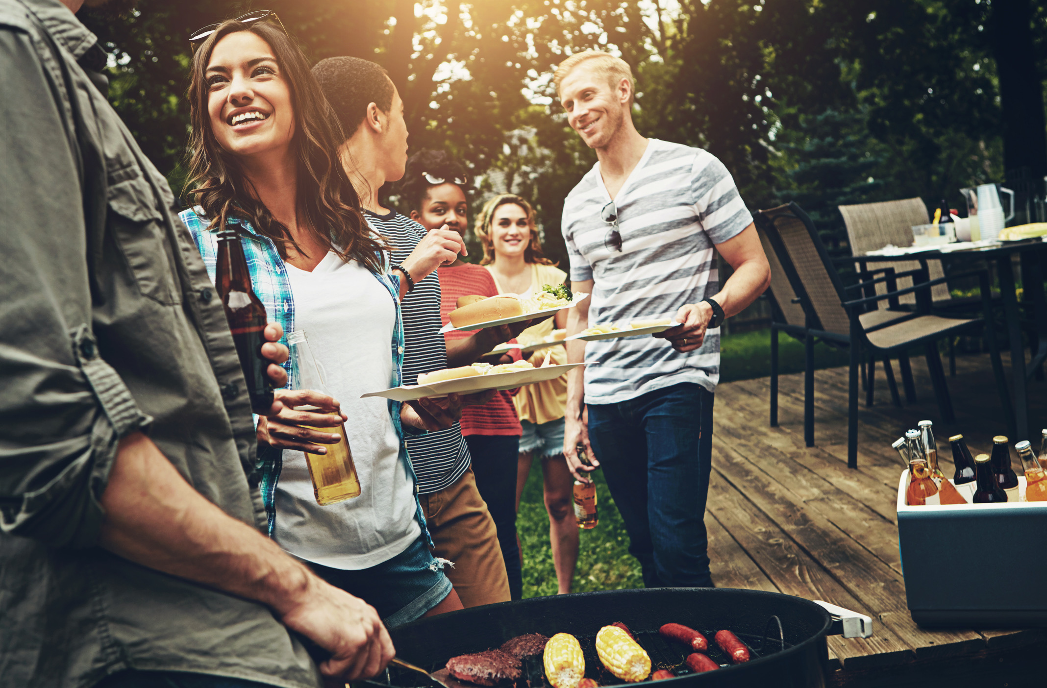 People enjoying a backyard barbecue, grilling hot dogs, corn on the cob, and sausages on a grill surrounded by trees.