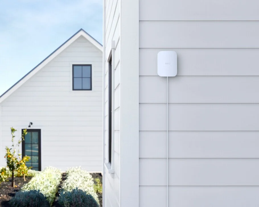 Close-up of a white exterior wall of a house with a small window, and a white electronic device mounted on the wall connected by a thin white wire.