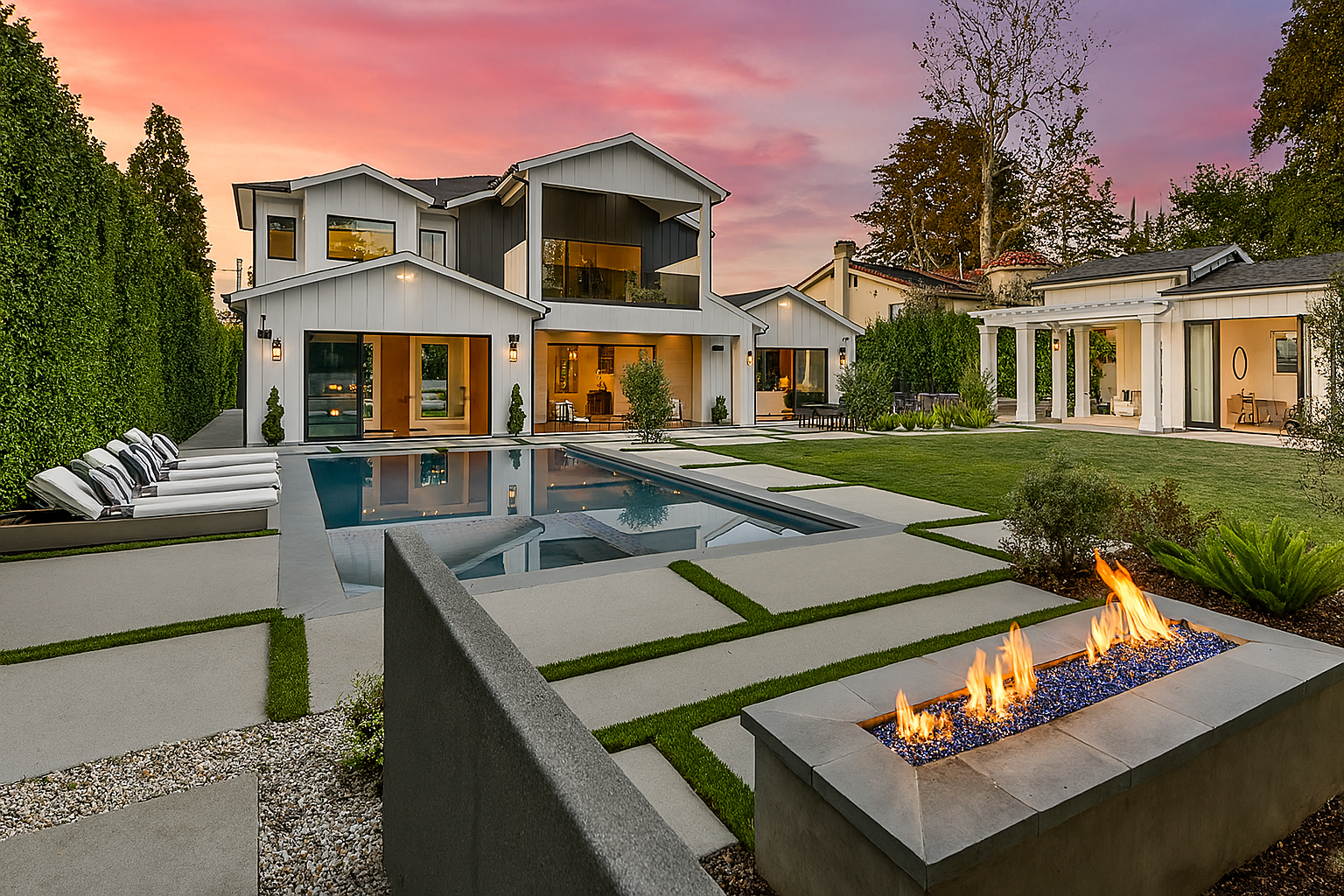 Modern backyard with a swimming pool, outdoor seating, and a fire feature, in front of a multi-story contemporary house during sunset.