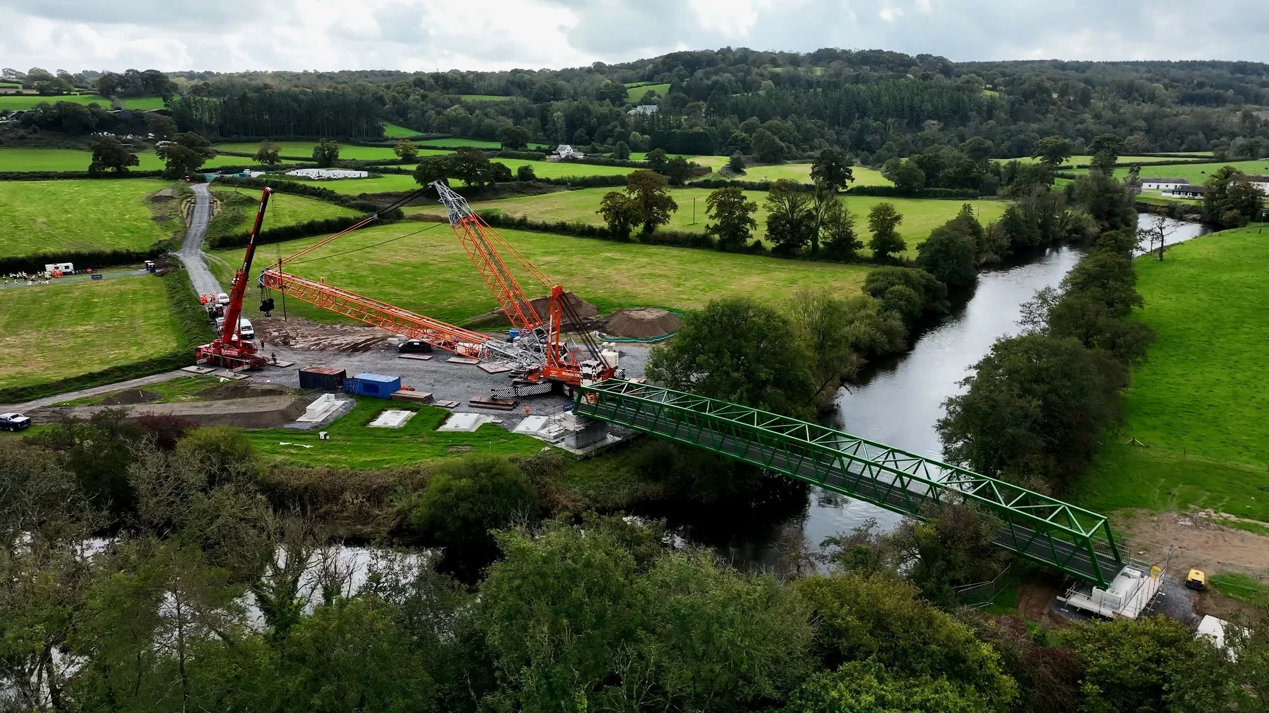 An aerial view of a construction site with cranes installing a bridge over a river in a rural area with green fields and trees.