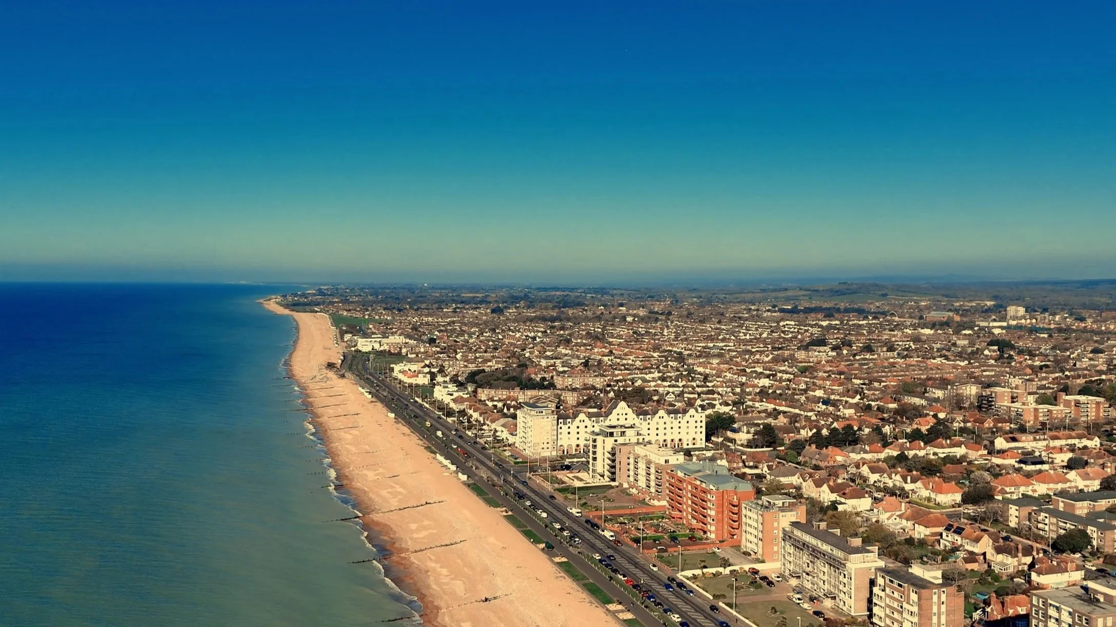 Aerial view of a beachside city with a sandy shoreline, a coastal road, and numerous buildings, with clear skies overhead.