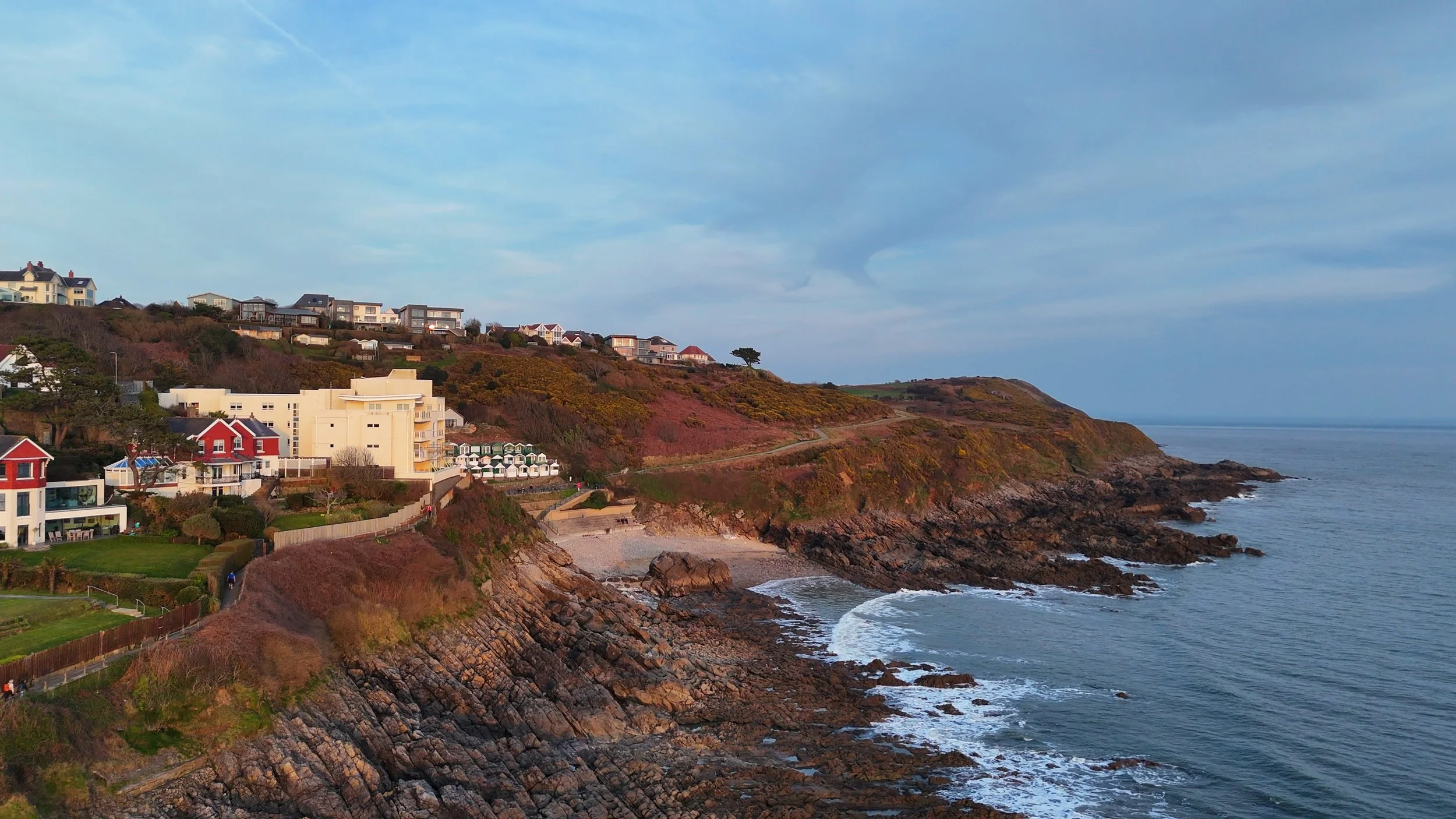 Coastal scene with rocky cliffs and a small beach, houses on the hillside, and the ocean extending to the horizon under a partly cloudy sky.