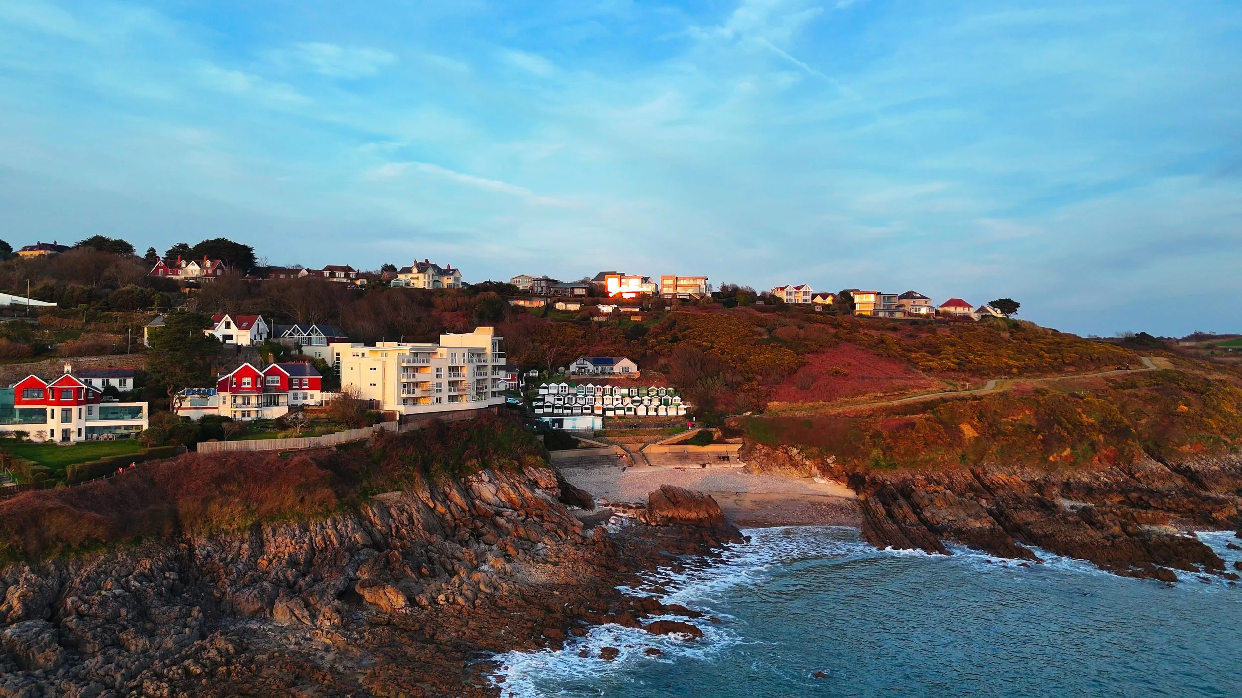 Scenic coastal view with houses on a hillside and rocky shoreline at sunset.
