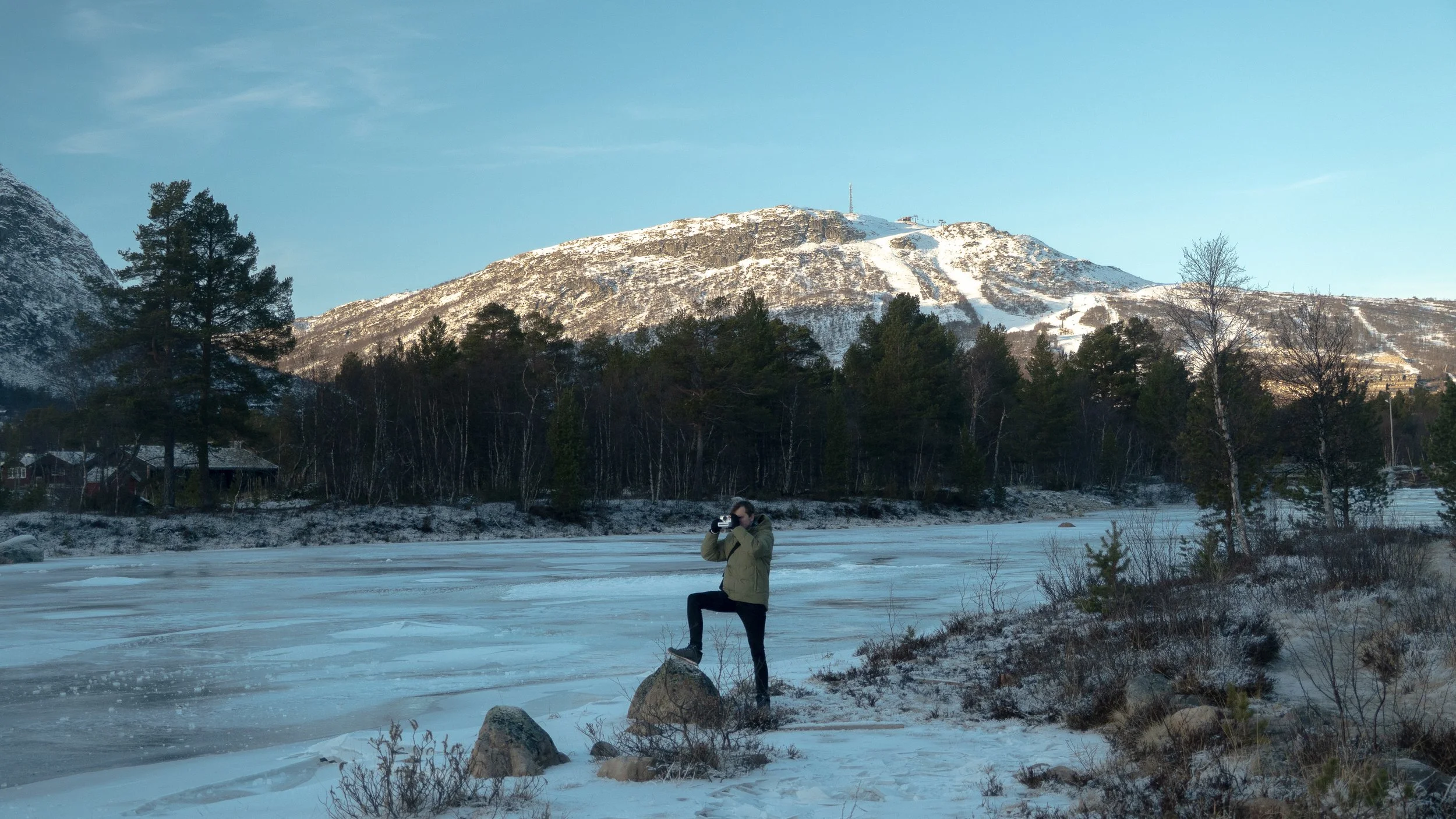 A person standing on a snowy riverbank taking a photograph with a camera, with snow-covered mountains and pine trees in the background.