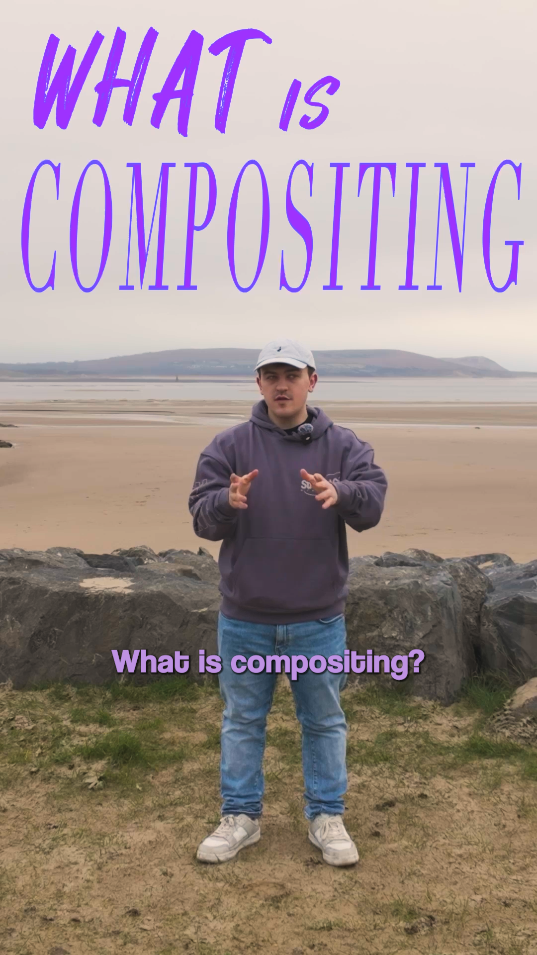A young man standing on a rocky beach with a body of water and distant hills in the background, wearing a gray hoodie, jeans, and a baseball cap, with text overlay asking 'What is composting?' and 'What is composting?' in purple and pink font.