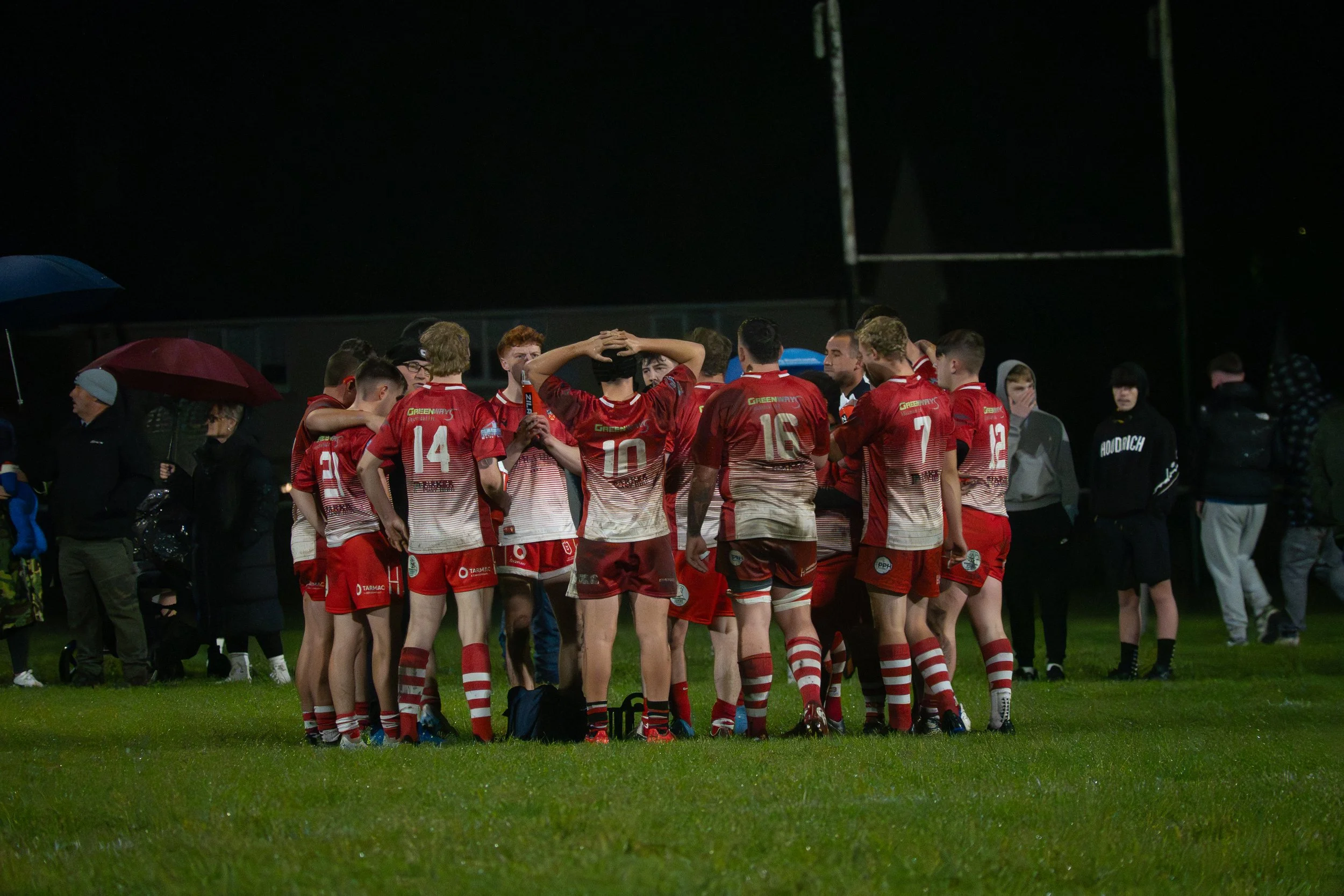 A group of rugby players in red and white uniforms gathered together on a field during a game at night, listening to their coach.