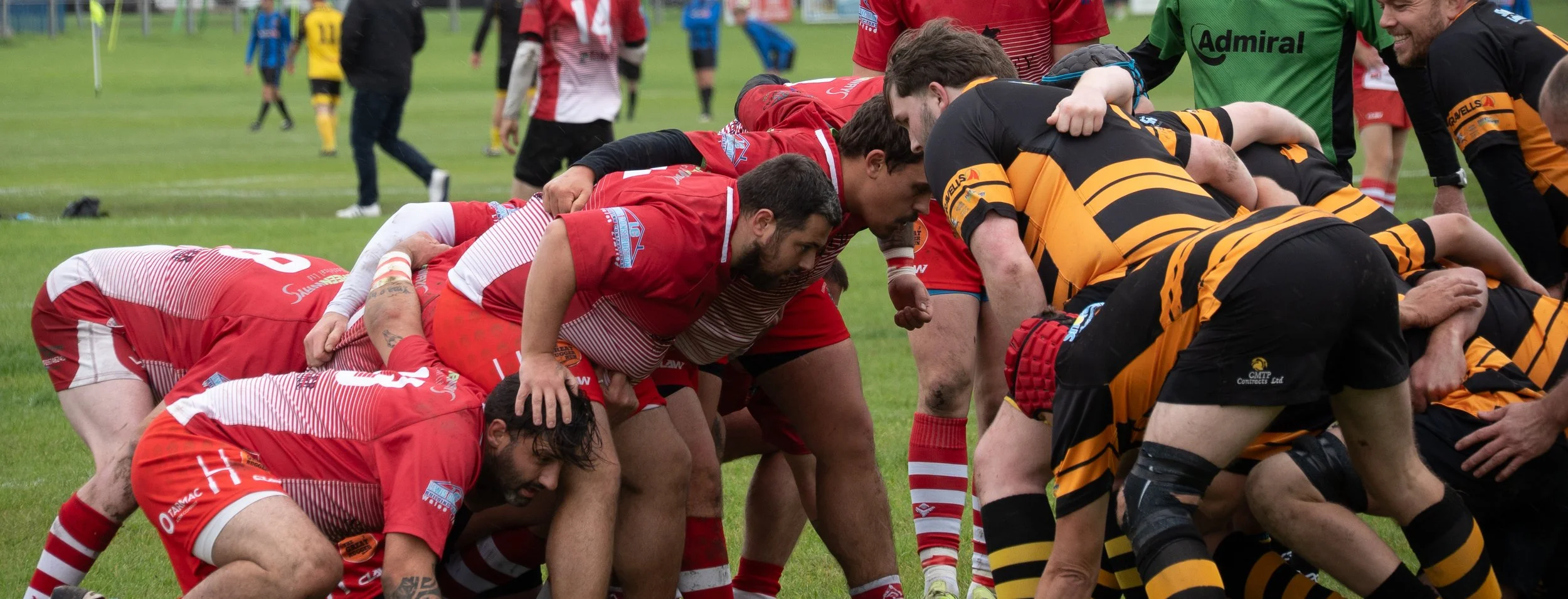 Rugby players in a scrum during a match, with players in red and black uniforms facing each other on a grassy field.
