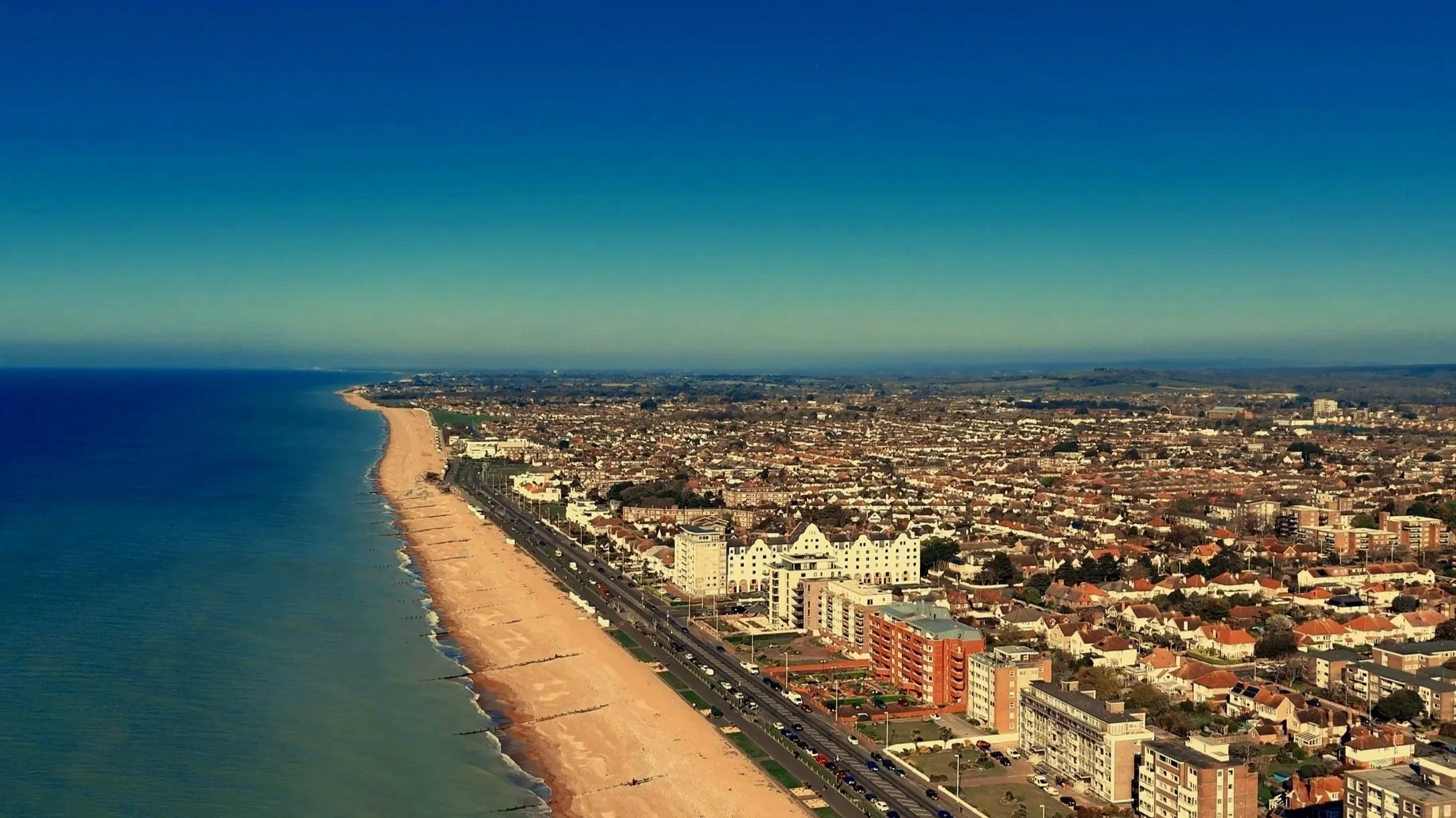 Aerial view of a coastal city with a long sandy beach and ocean on the left, and a densely populated urban area with various buildings and streets on the right.