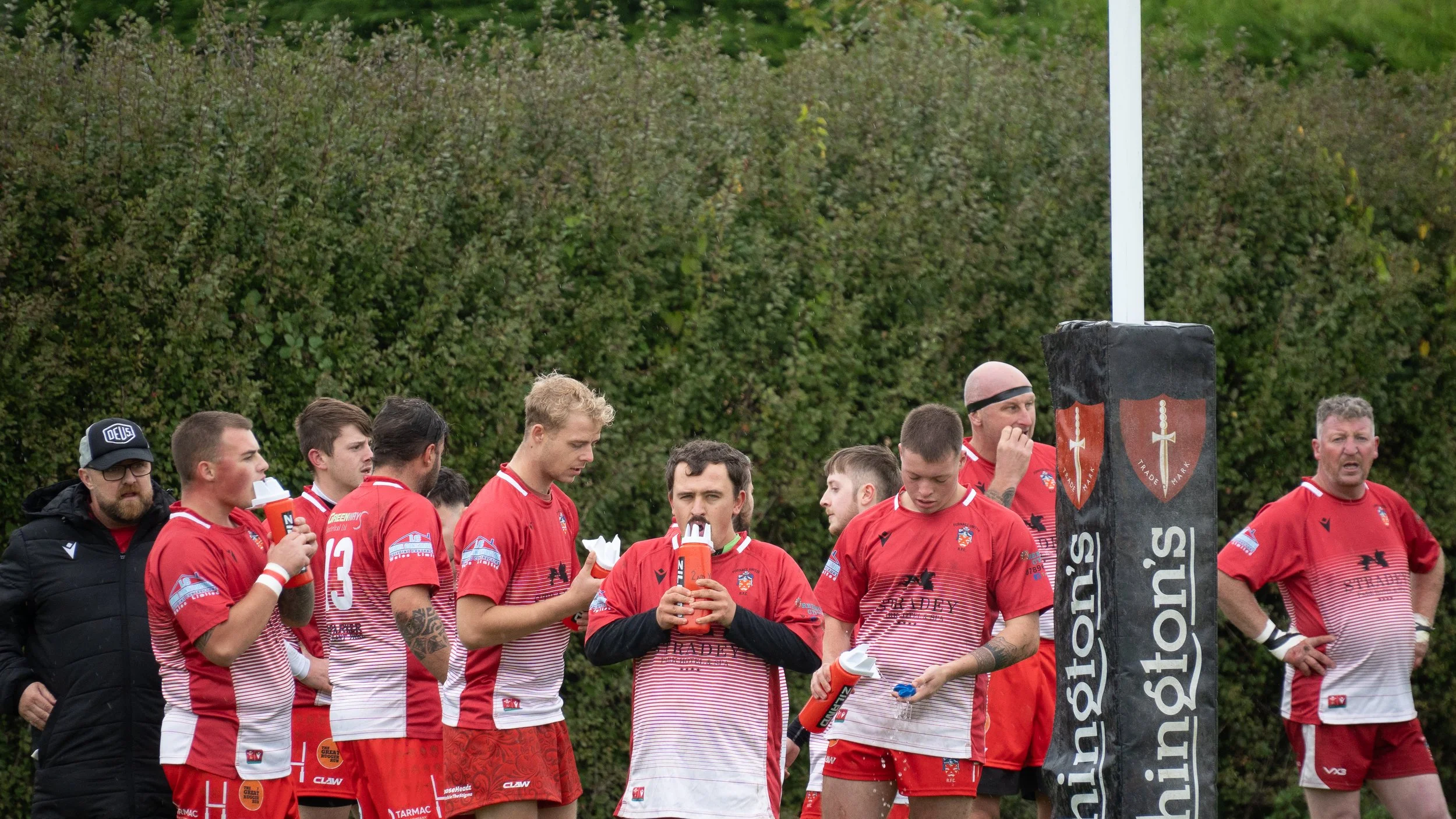Rugby players wearing red and white uniforms standing on a field, some drinking water, with a coach or team staff on the left, in front of a green shrubbery background. A padded rugby goalpost with branding is also visible.