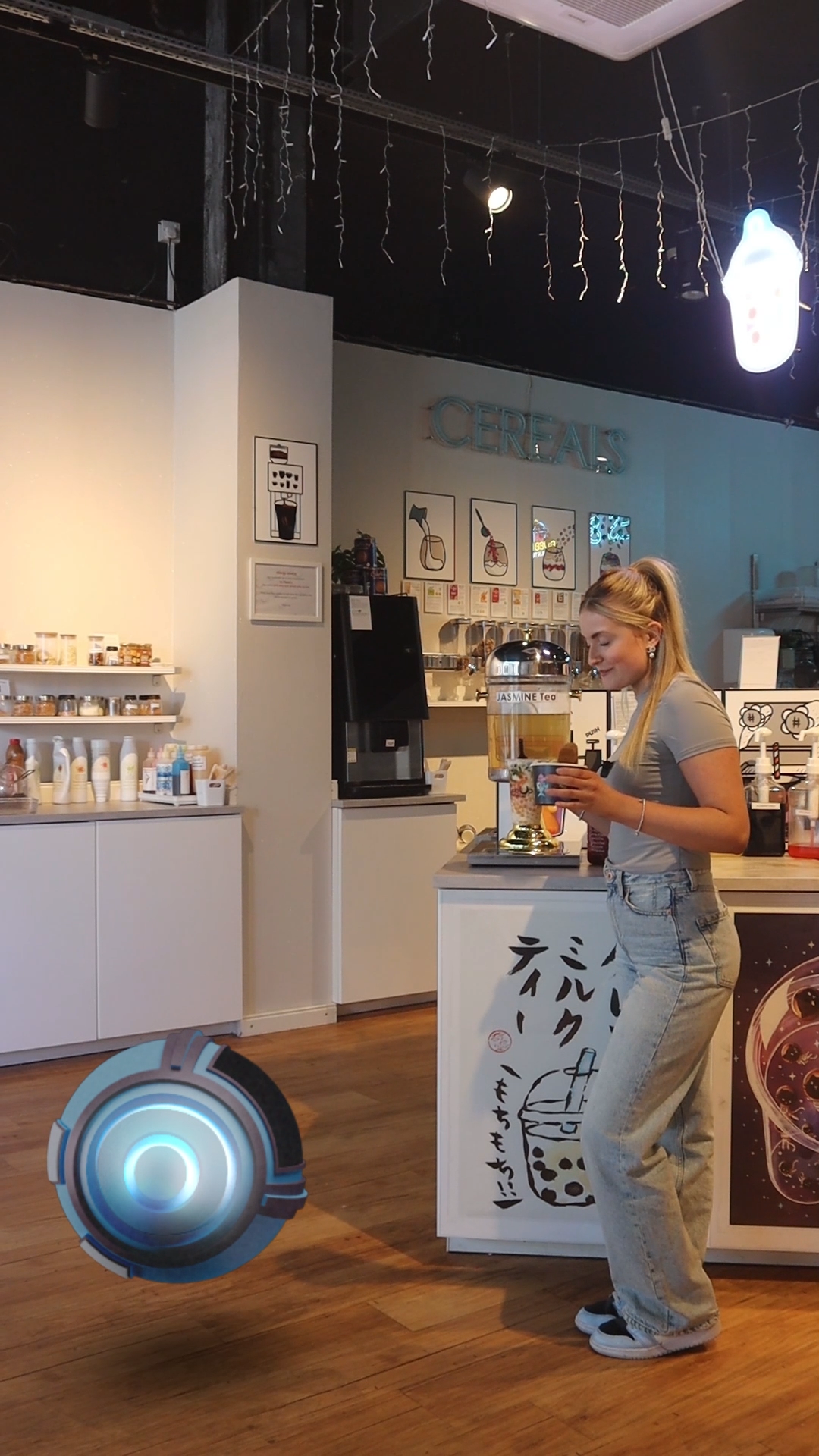 A young woman in casual clothing is standing at a bubble tea shop, holding a cup of tea with tapioca pearls, with a digital robot or drone hovering near her. The shop has a modern interior with white shelving, boxes, and a counter labeled with Japanese characters and an illustration of a bubble tea cup.