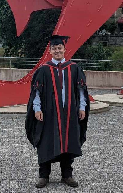 Graduation cap and gown worn by a young man standing outdoors in front of a red sculpture.