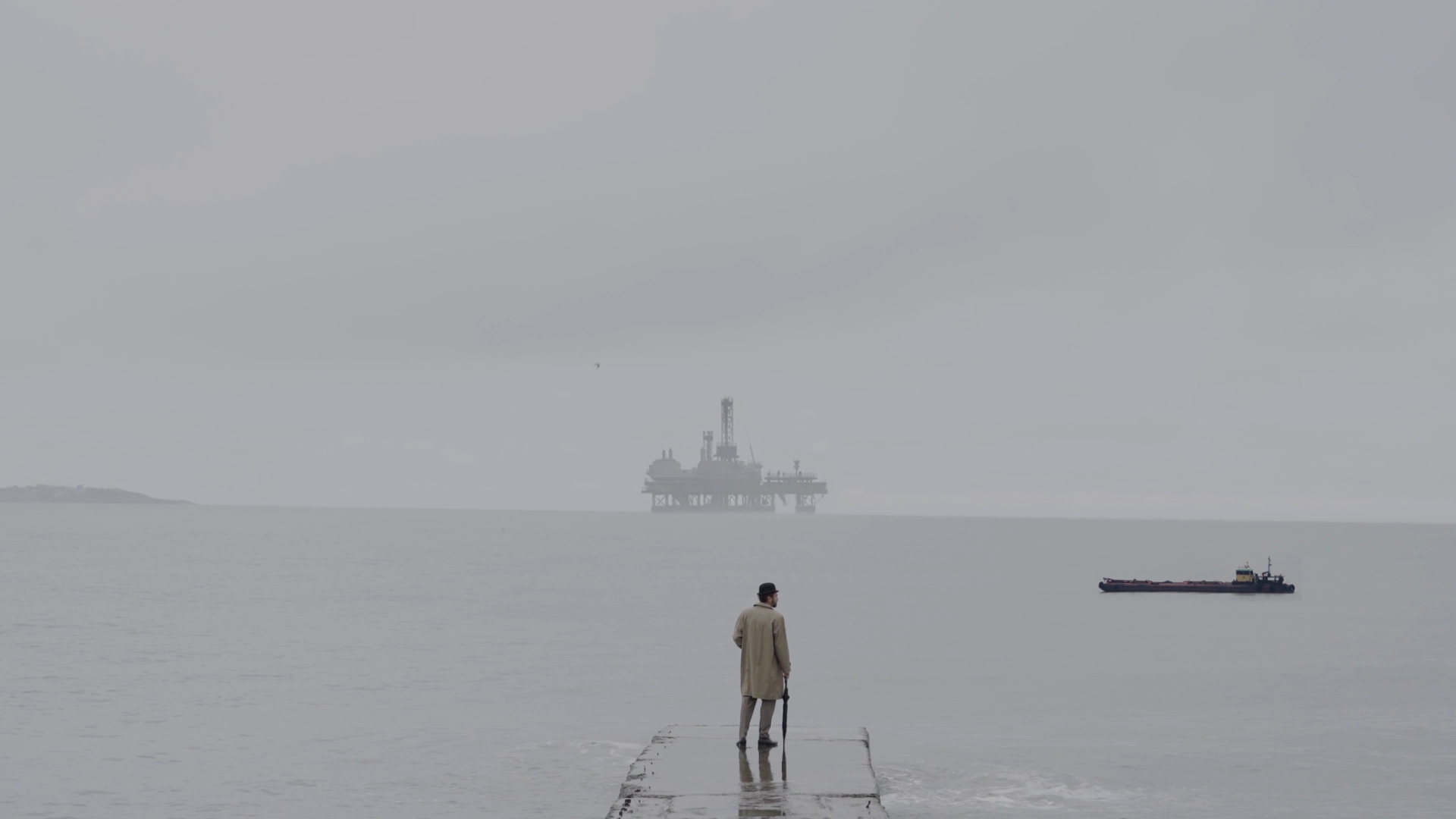 An older man standing on a narrow pier by the water, holding an umbrella, looking at an offshore oil rig in the distance on a cloudy day.