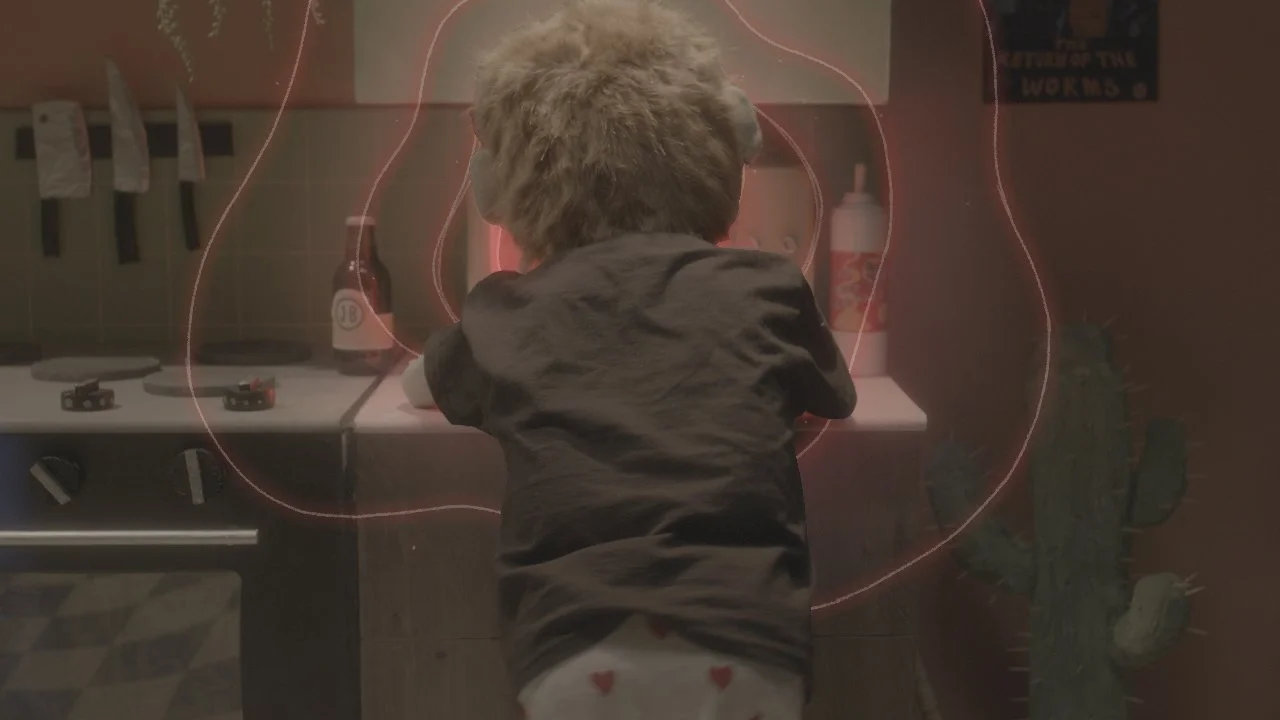 A young child with curly hair wearing a dark shirt and light-colored underwear with hearts, seen from behind, playing with a toy kitchen set that includes bottles and play food, in a kitchen with a cactus plant nearby.