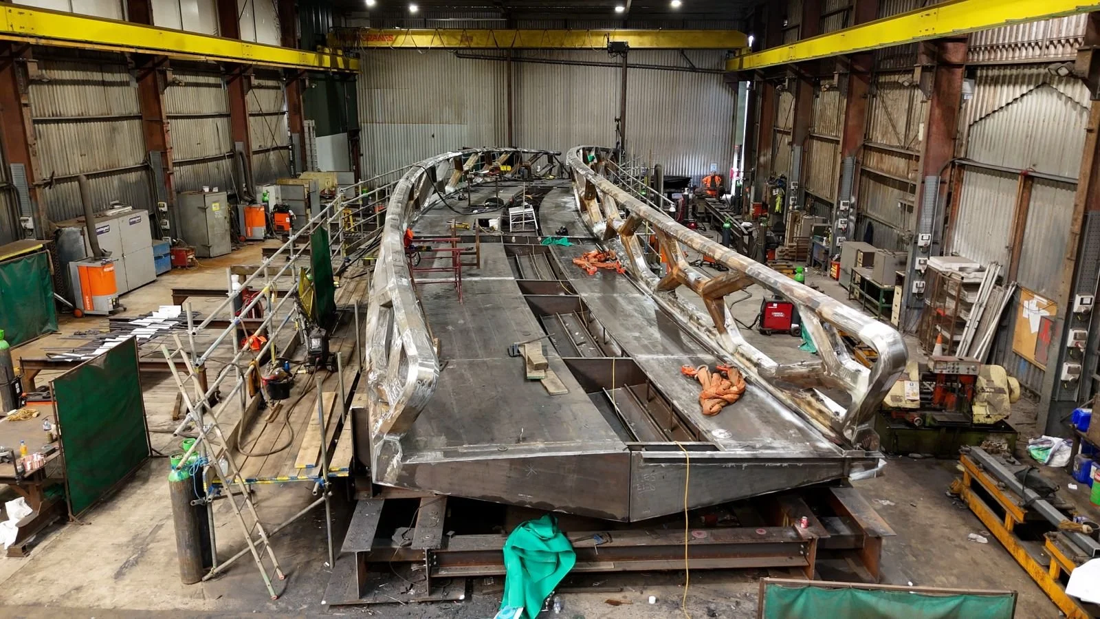 An industrial workshop with a large steel structure of a boat being assembled, surrounded by tools, machinery, and workers in protective gear.