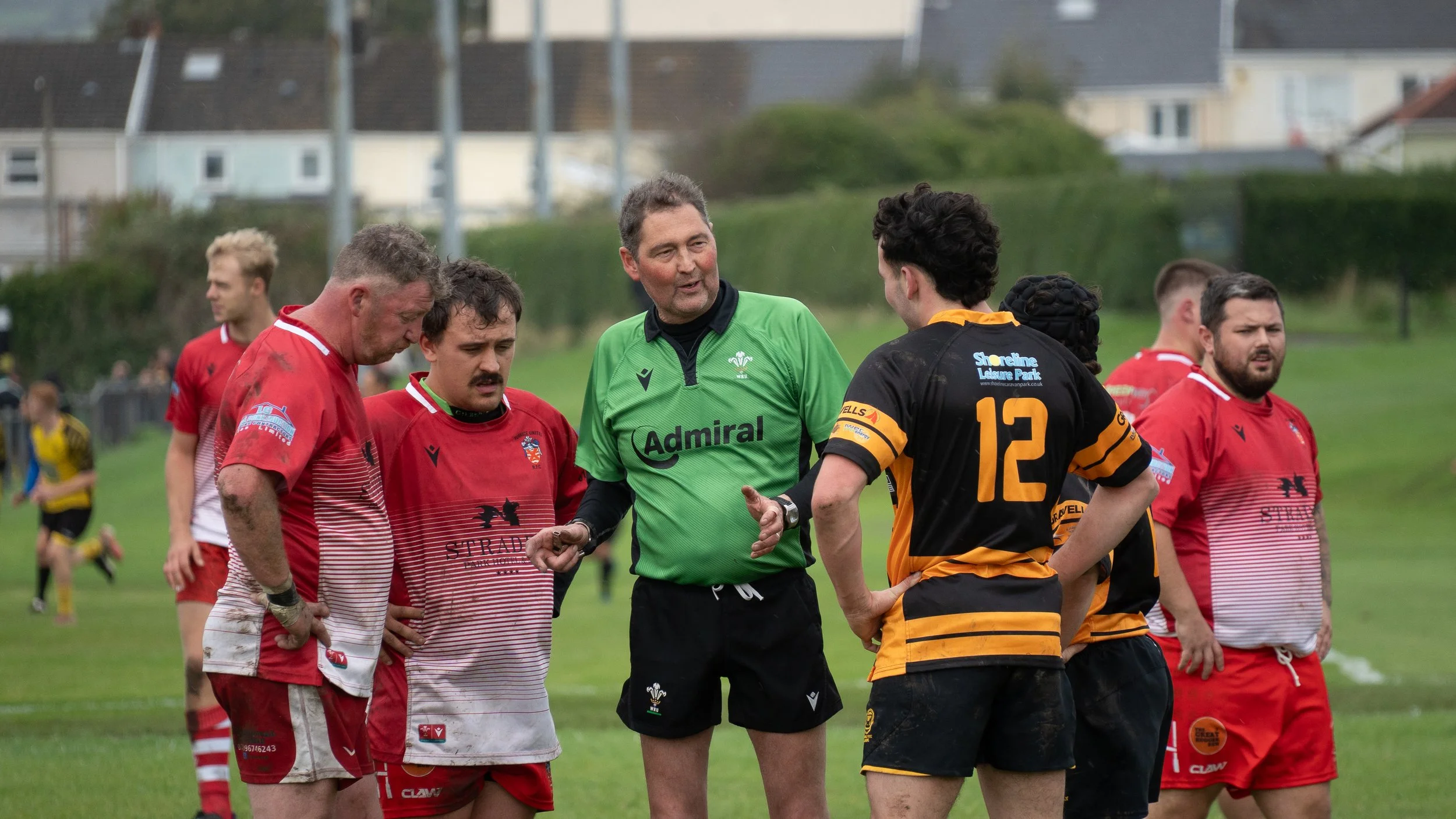 Rugby players and referee talking on the field before a match, with players in red and black uniforms and the referee in green.