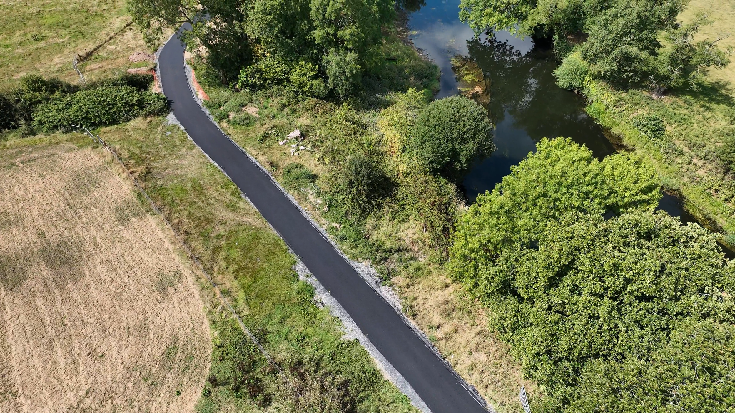 Aerial view of a rural landscape showing a newly paved winding road running alongside a pond with green trees and grass on either side.