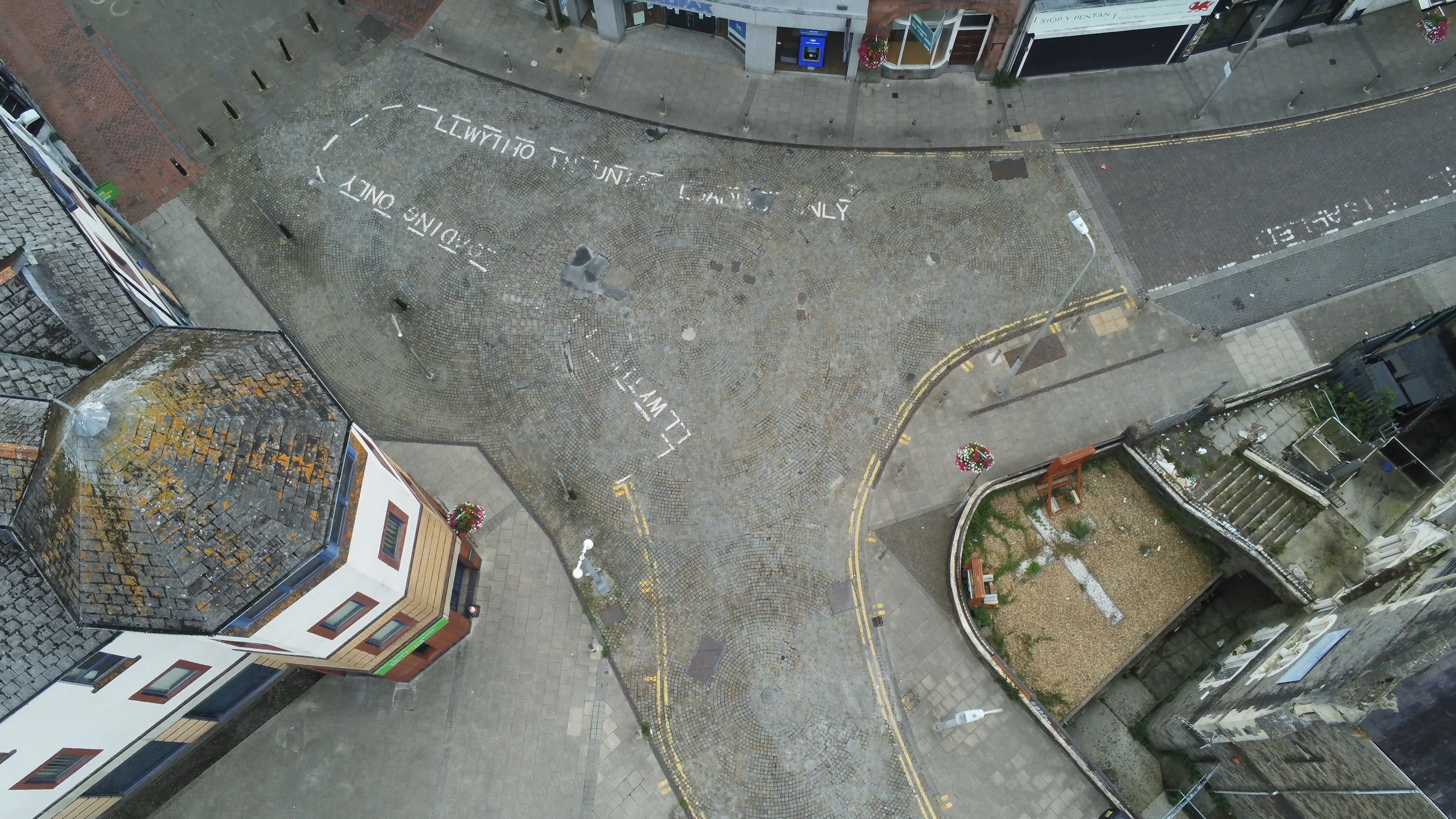 An aerial view of a cobblestone street with painted parking and traffic lane markings, surrounded by buildings and small sidewalk areas with street lamps and flower pots.