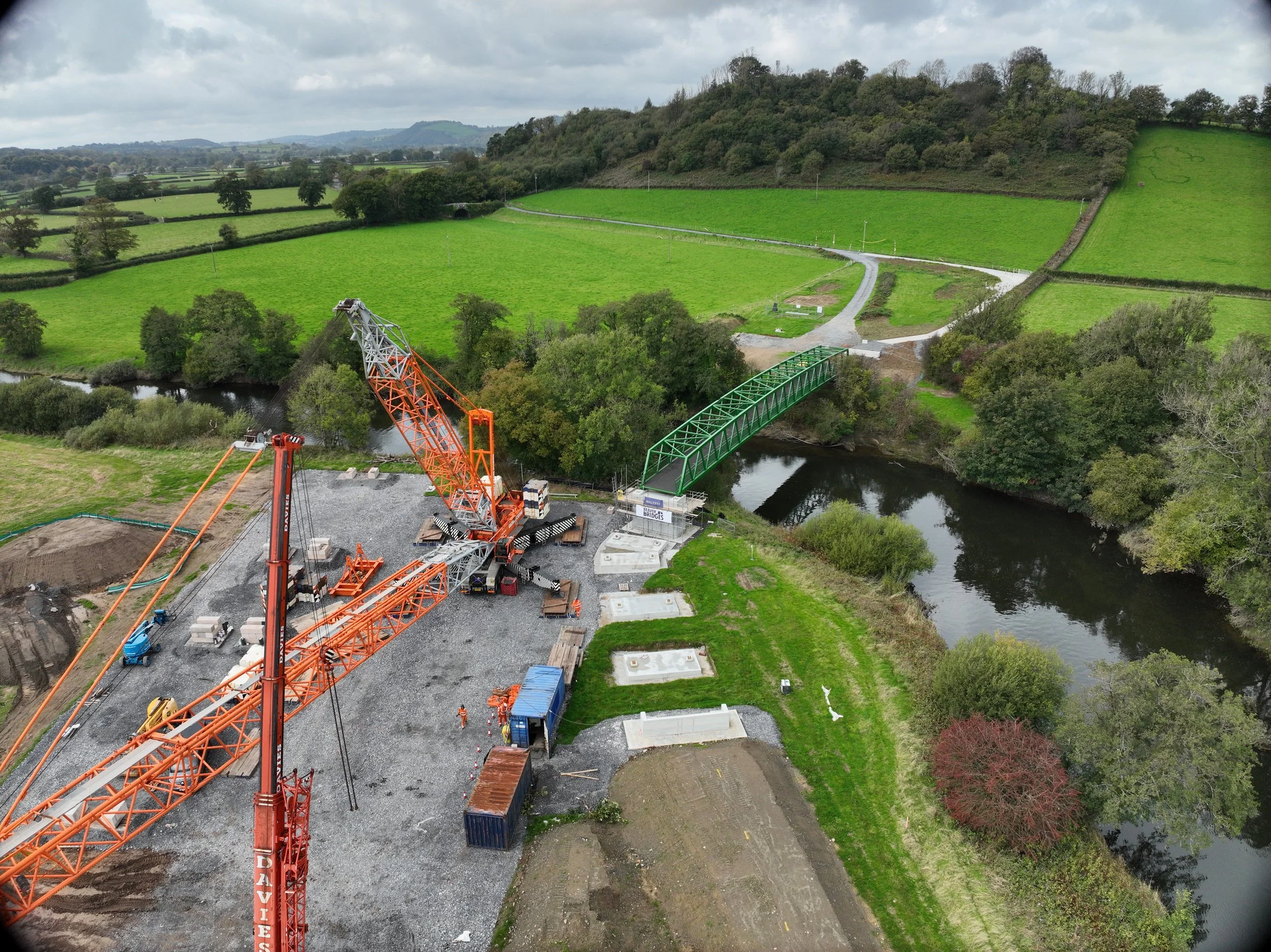 A construction site with a large orange crane next to a small river, as a green metal bridge is being built over the water amidst a rural landscape with green fields and wooded hills.