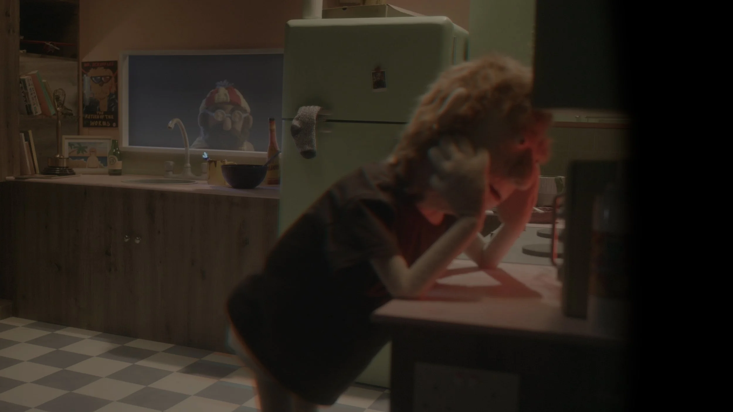 A young boy with curly hair appears upset, holding his head in his hands, in a dimly lit kitchen. In the background, a woman with curly hair is leaning over a counter, and a refrigerator with a small magnet is visible. A computer monitor on the counter shows an animated character with glasses and a hat.