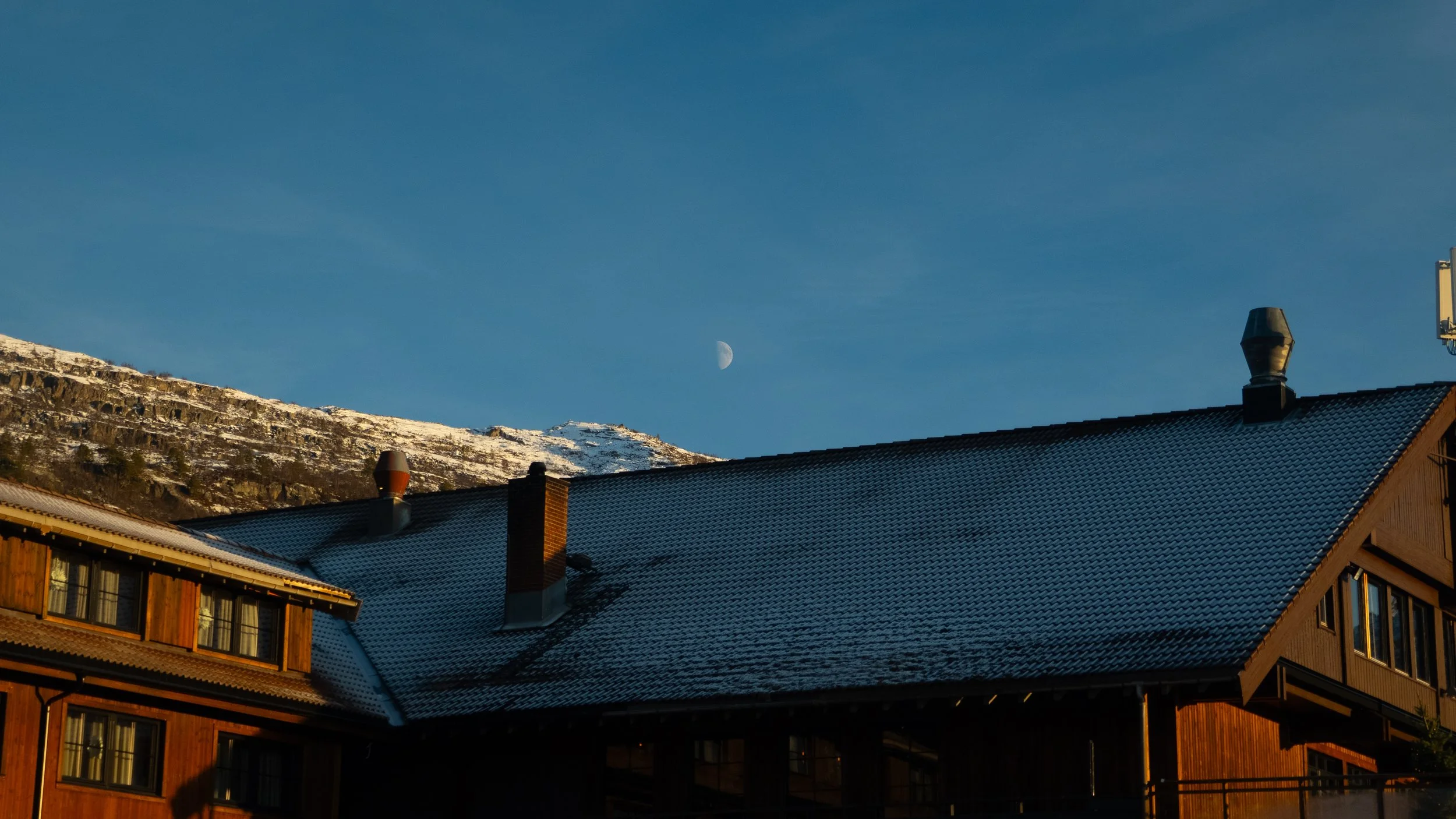 A house with a snow-covered metal roof, set against a mountain backdrop, with the moon visible in the clear blue sky.