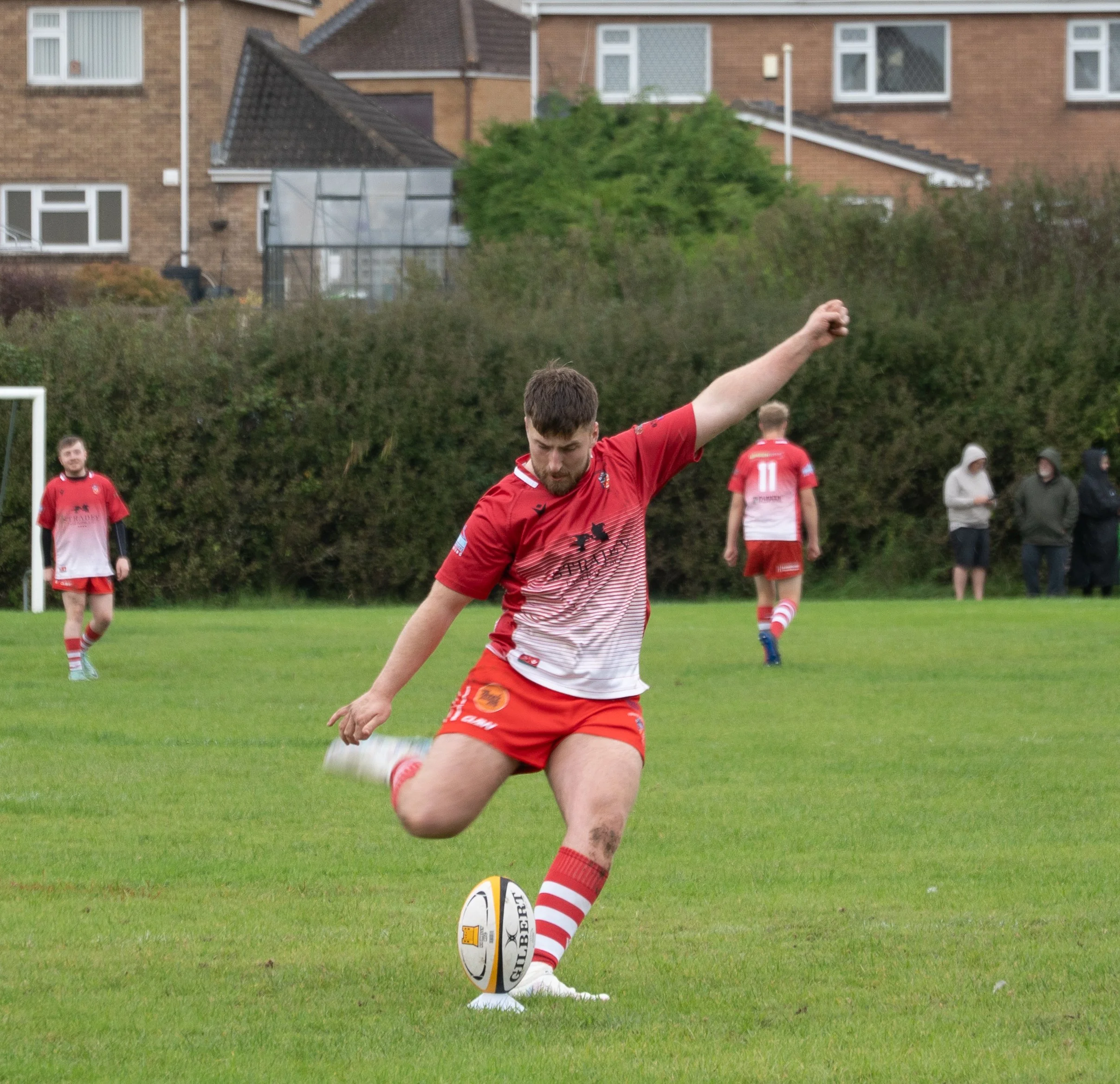 A rugby player in a red uniform kicks a rugby ball on a grassy field, with other players and spectators in the background.