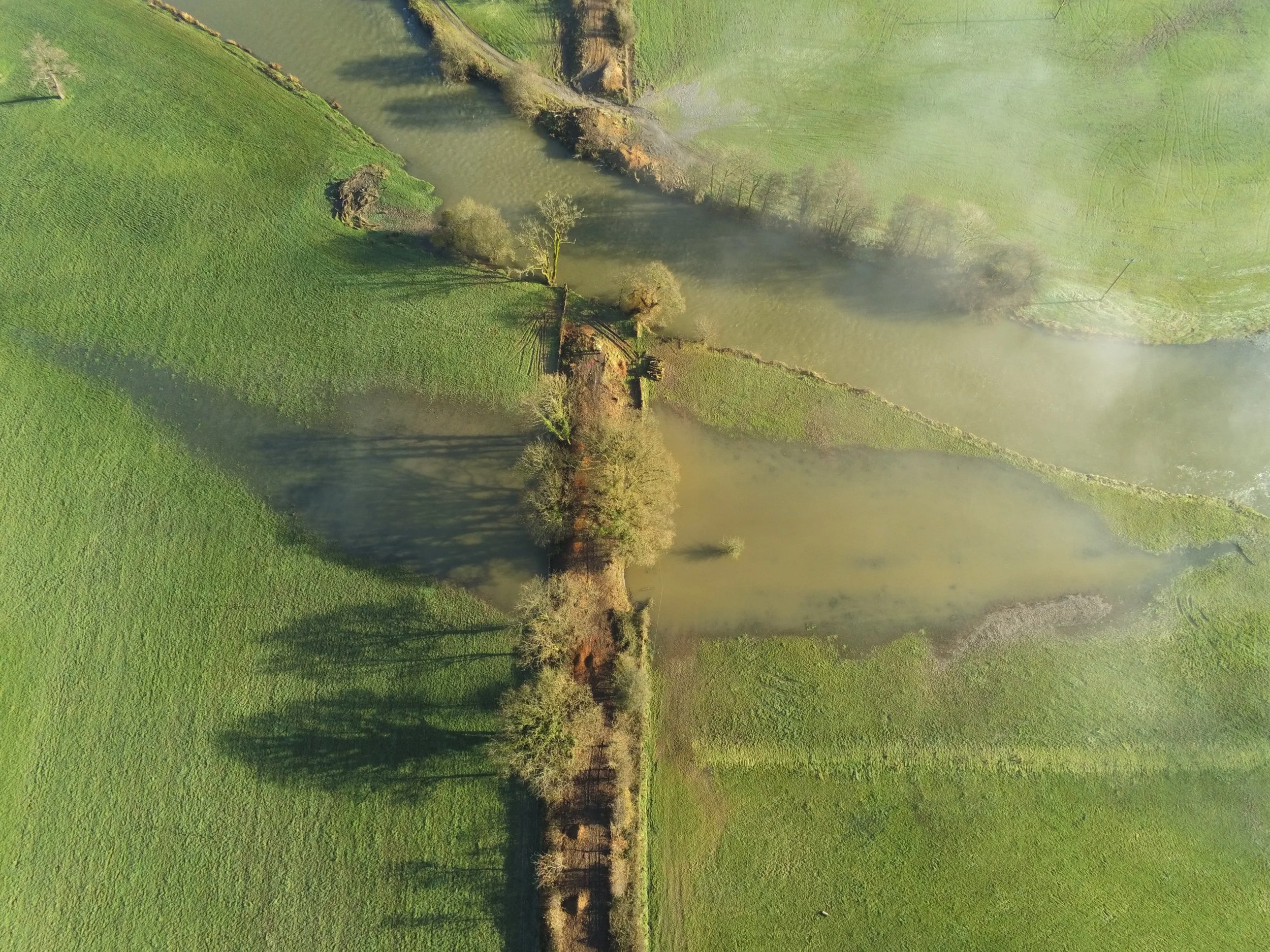 An aerial view of a flooded landscape with lush green fields, trees, and water bodies, featuring mist or fog near the water.