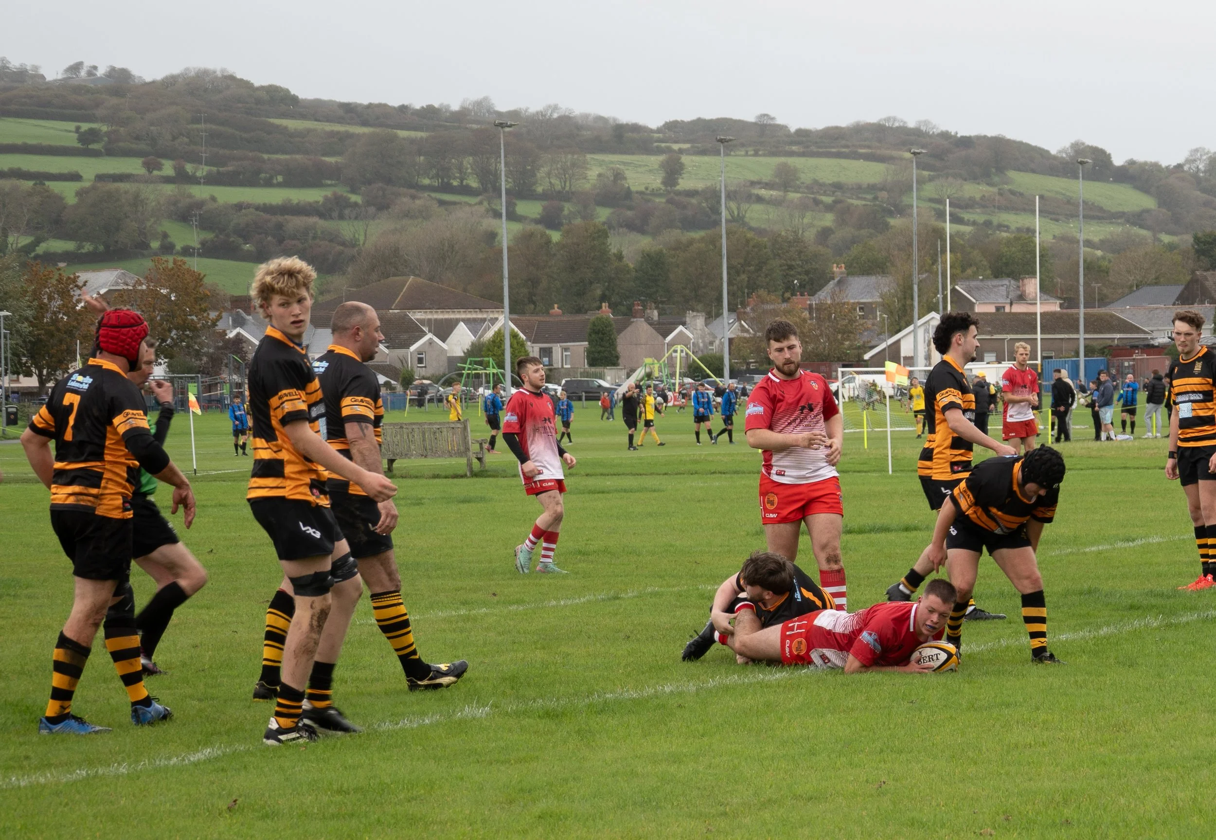 Rugby match on a grassy field with players in red and black and yellow stripes uniforms. One player in red is on the ground holding the ball, tackled by a player in black and yellow. Other players are standing nearby, watching or preparing for the next play, with a park and hills in the background.
