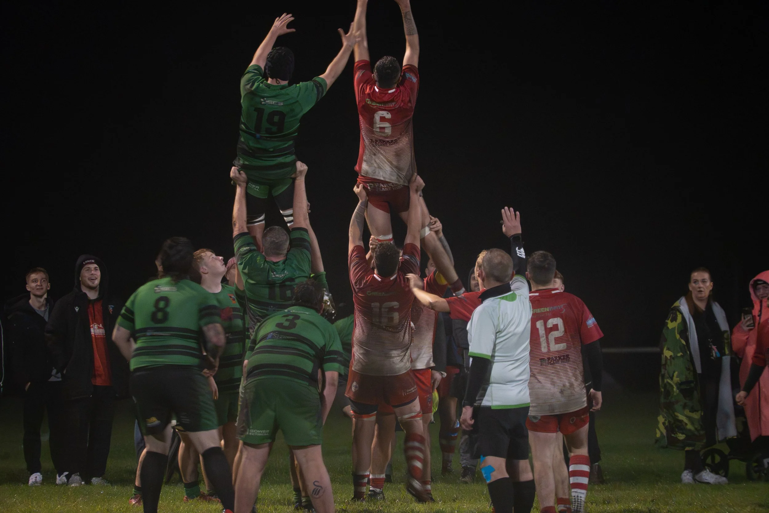 Rugby players forming a human pyramid during a match, with some players lifting others into the air, and there are onlookers in the background.