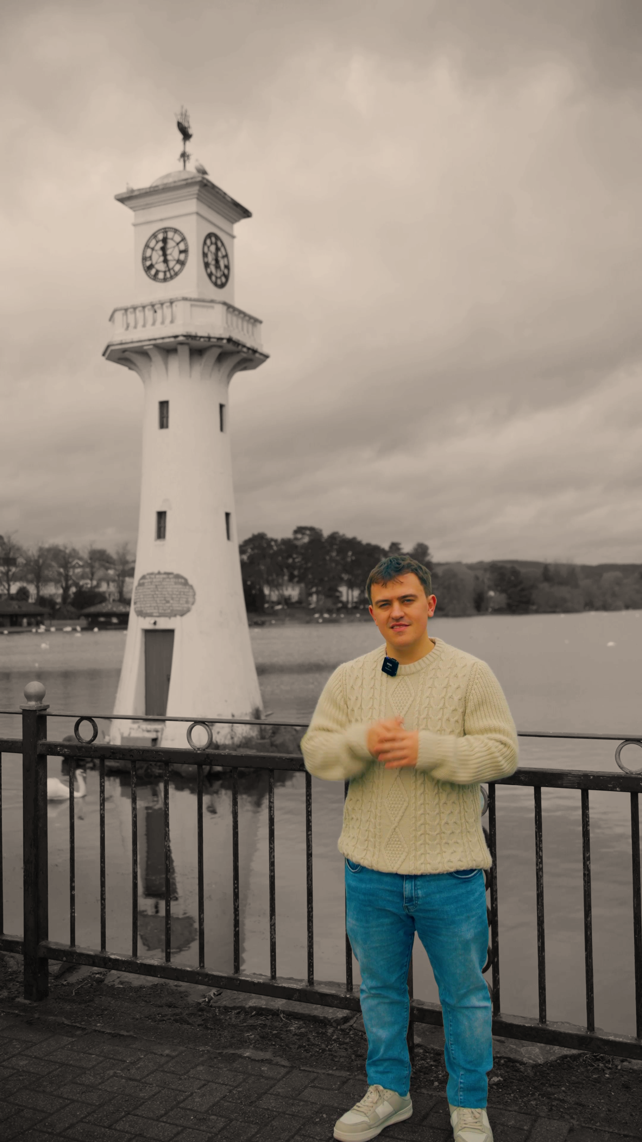 A young man standing by a black metal fence near a lake with a white clock tower in the background, outdoors on a cloudy day.