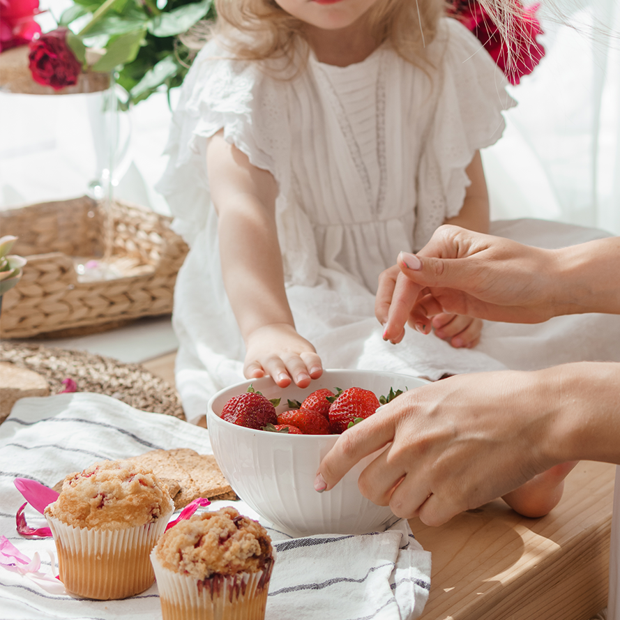 Child reaching for strawberries in a white bowl during a cozy indoor setting with baked goods in the foreground.