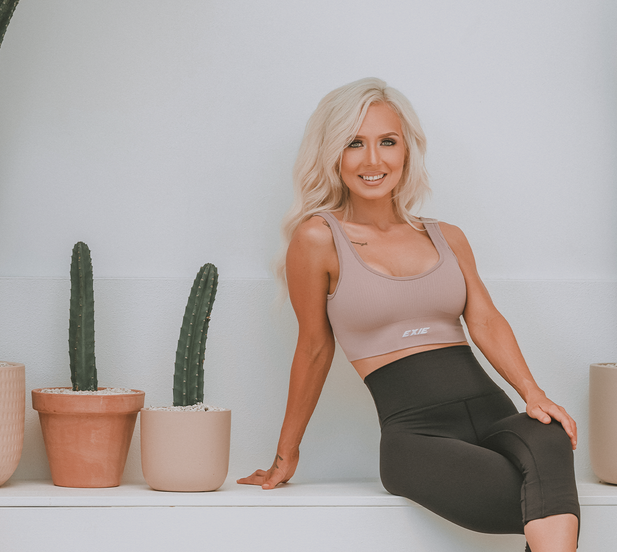 Bonnie Atkinson wearing a beige tank top and black leggings, sitting on a white surface with potted cacti against a plain white wall.