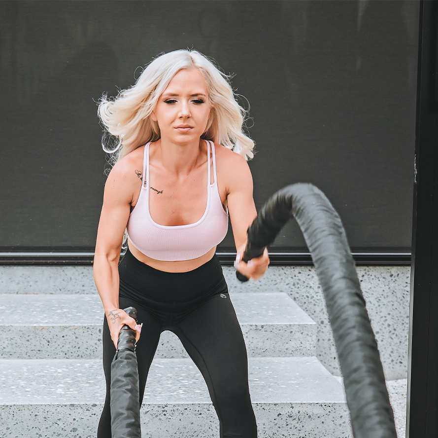 Bonnie Atkinson wearing a white sports bra and black leggings using battle ropes during workout outside.