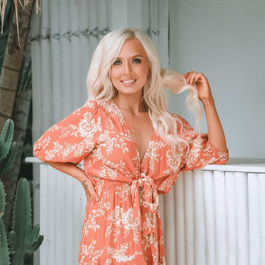 Bonnie Atkinson smiling, wearing a coral-colored floral dress with puff sleeves and a waist tie, standing indoors next to a white textured wall and green plants, including a cactus.